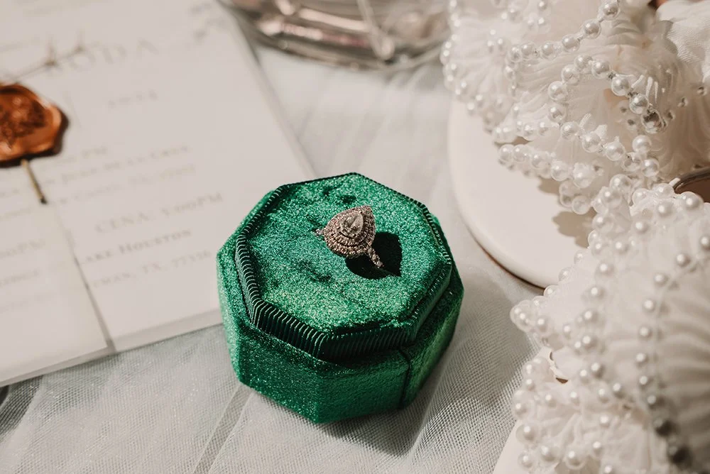 A ring in a green velvet jewelry box on a white surface surrounded by wedding or decorative items.