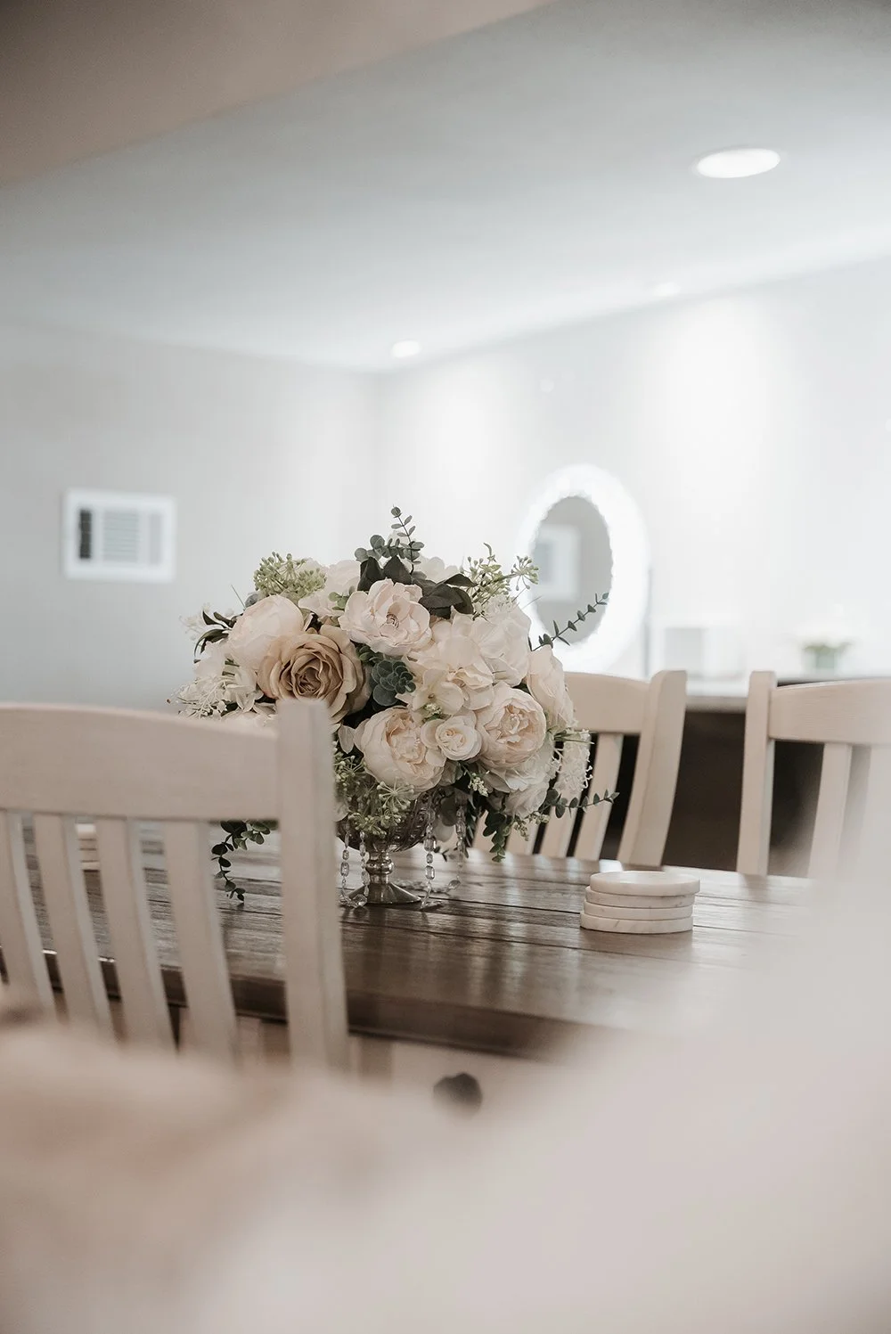 Elegant floral arrangement of white and beige roses and greenery on a wooden dining table in a bright room.