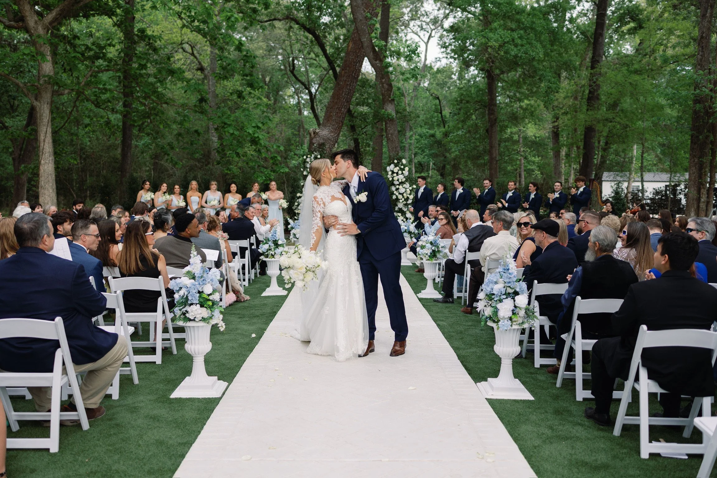 A bride and groom share a kiss during their outdoor wedding ceremony, with guests seated on white chairs and a backdrop of tall trees and floral arrangements.