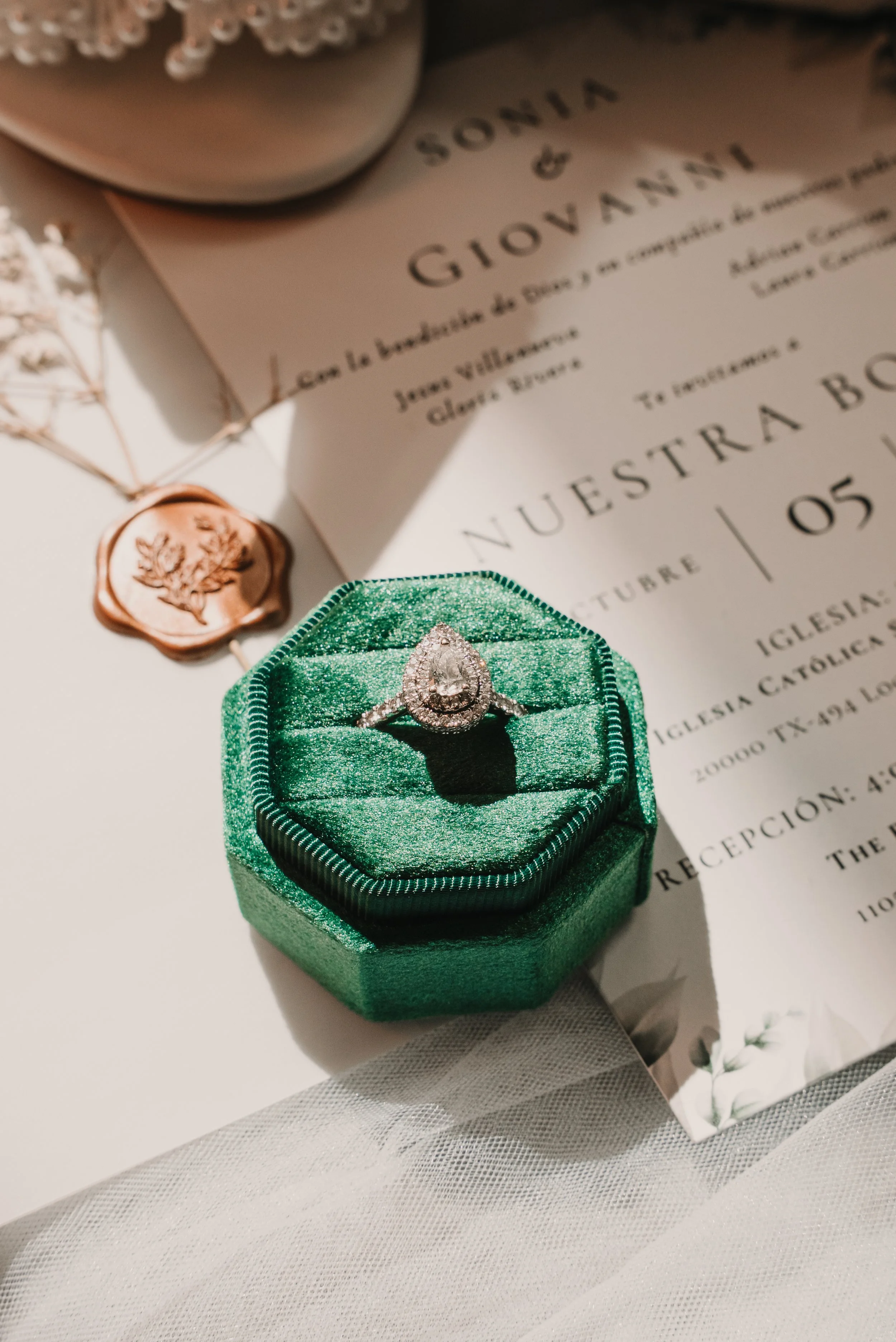 A diamond engagement ring in a green velvet ring box, a wedding invitation, and a wax seal are arranged on white fabric.