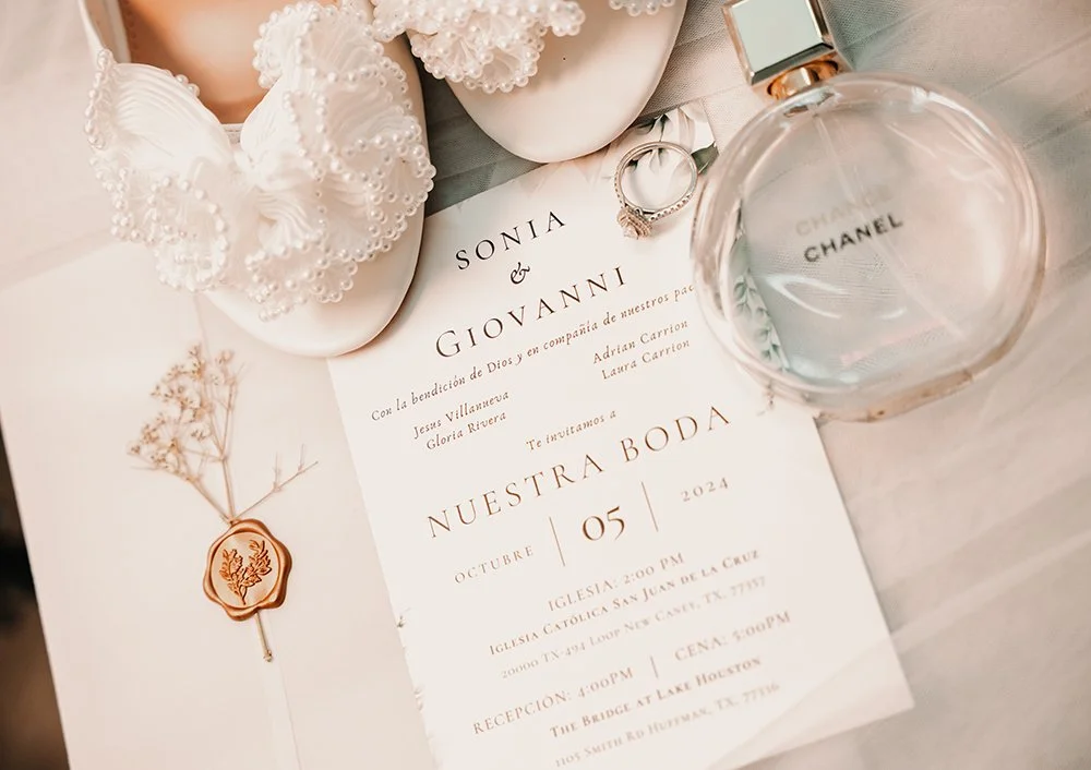 Wedding invitation, baby shoes with pearl beads, a bottle of Chanel perfume, an engagement ring, and a small sprig of dried flowers with a wax seal, all placed on a white surface.