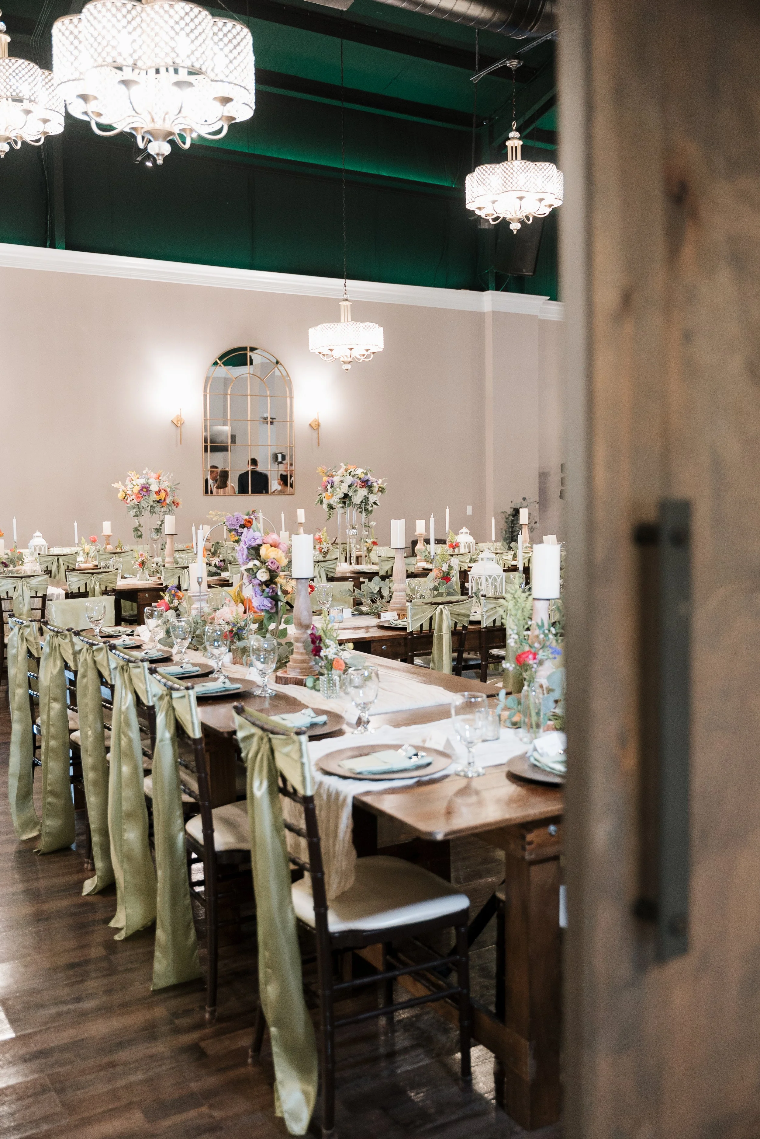 A banquet hall decorated for a special event with long tables, floral centerpieces, and candles. Elegant chandeliers hang from the ceiling, and there is a large mirror on the wall.