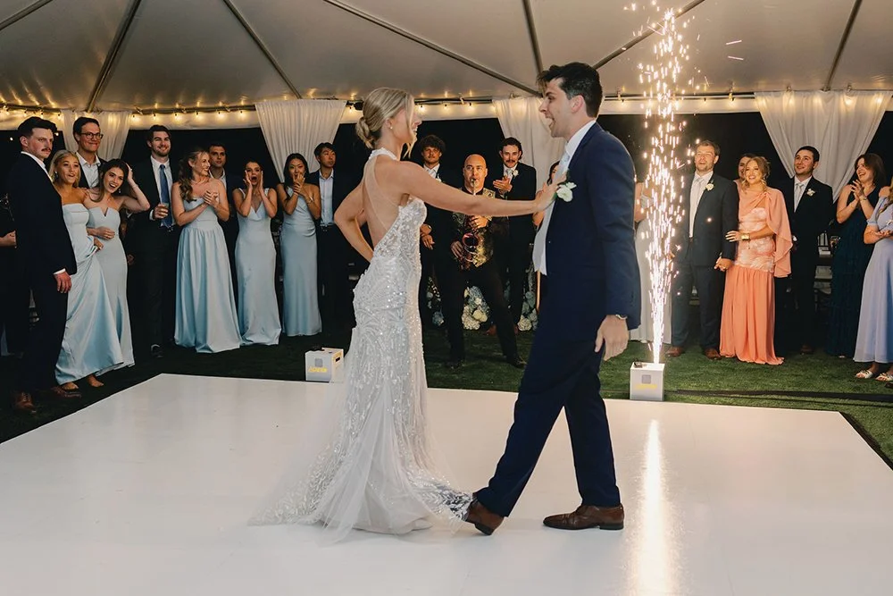 Bride and groom dancing at their wedding reception with guests watching in a tent with string lights, and fireworks in the background.