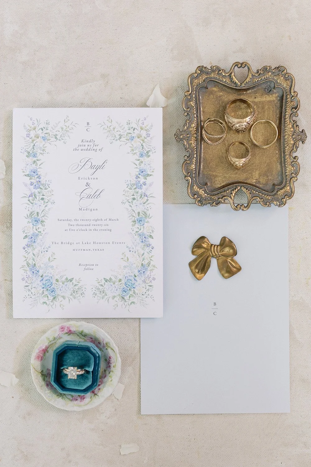 Wedding invitation, rings on a tray, gold bow decoration, and engagement ring in a box.