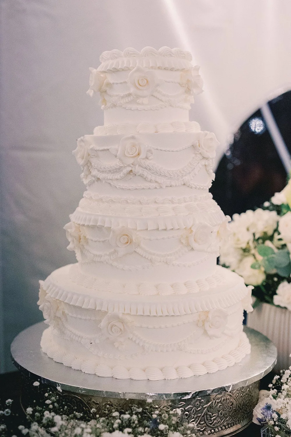 A tall, white, multi-tiered wedding cake decorated with piped icing and roses, placed on a decorative silver cake stand surrounded by white flowers.