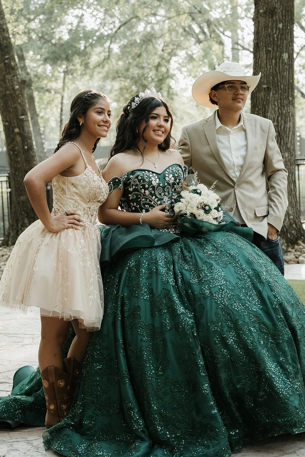 Three people, including a bride in a green gown holding a bouquet of white roses, posing outdoors with trees in the background.
