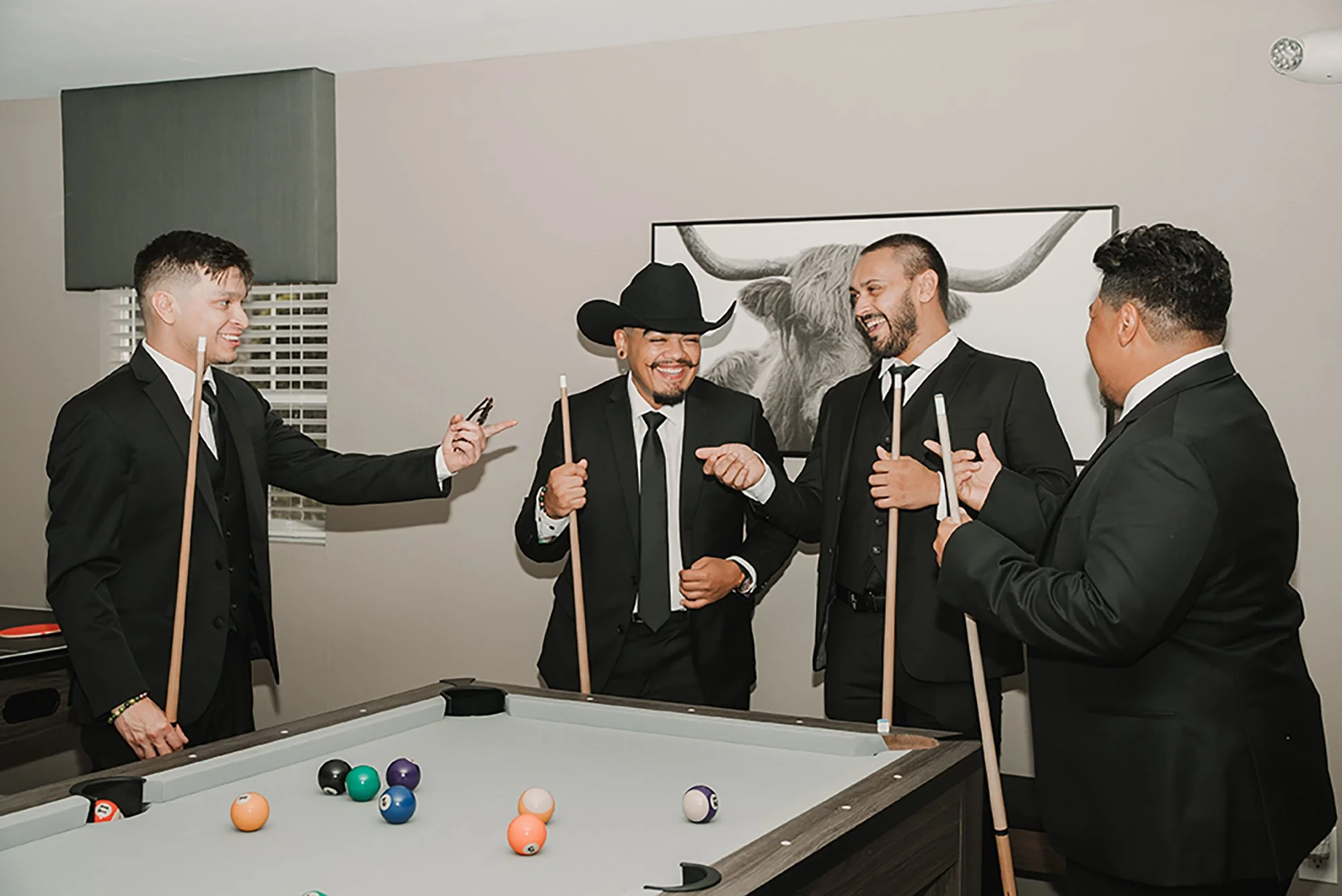 Four men dressed in black suits and ties are playing pool in a room with a pool table. One of the men is wearing a cowboy hat and they are all smiling, enjoying their game and conversation. Billiard balls are spread out on the table, and there's a large framed picture of a longhorn steer hanging on the wall behind them.