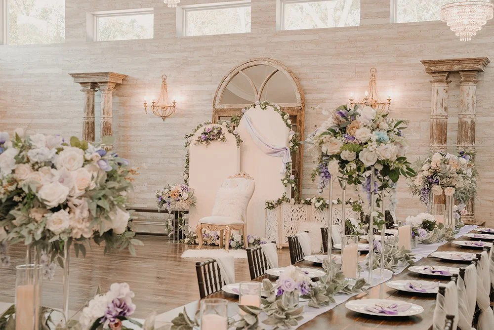 Elegant indoor wedding reception with floral centerpieces, candles, and a decorated backdrop with a chair for the bride or couple.