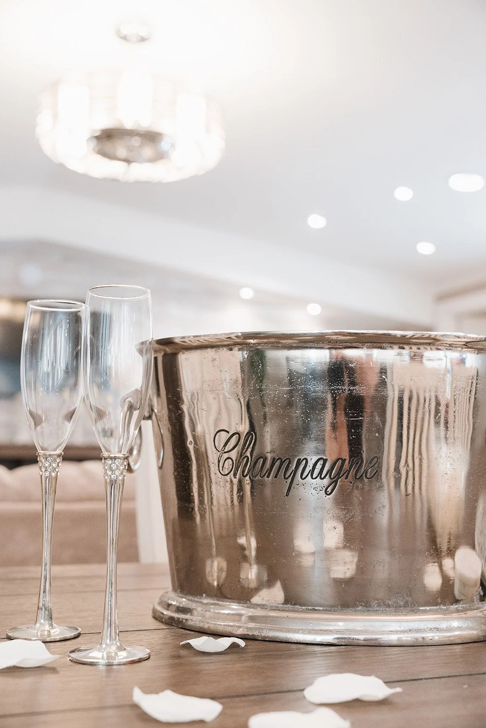 Silver champagne bucket labeled 'Champagne' with two empty champagne glasses beside it on a wooden table, surrounded by scattered white rose petals.