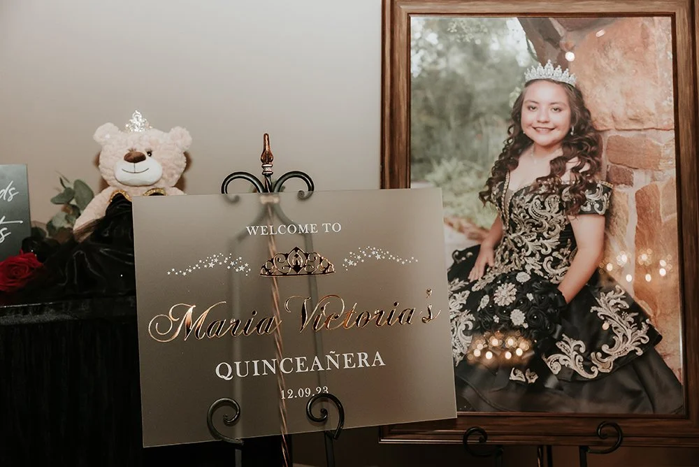 A framed photo of a young girl in a fancy black and gold dress with floral patterns, sitting outdoors against a stone wall, wearing a tiara and smiling. Next to it is a welcome sign for Maria Victoria's quinceañera event, decorated with stars and a tiara illustration, and a teddy bear wearing a tiara and a black cape.