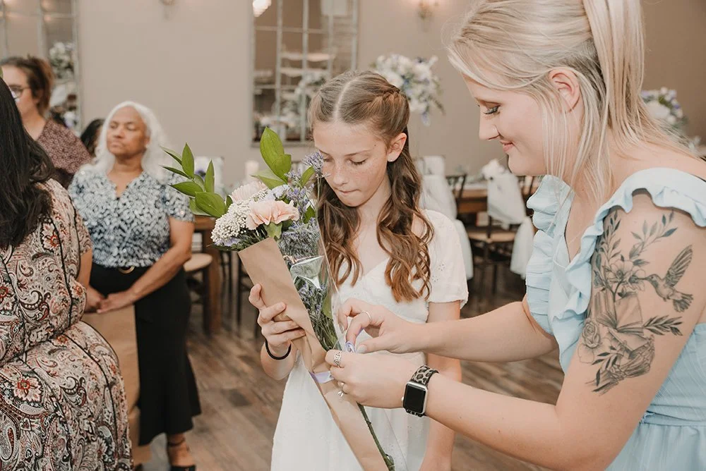 A woman with tattoos on her arm giving a flower to a young girl at a gathering, with other women in the background.