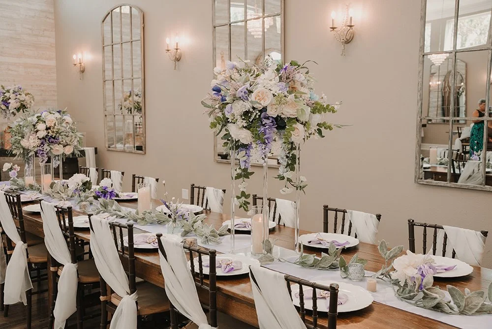 Long wooden dining table decorated with tall flower arrangements and candles, set for a formal event in a well-lit room with mirrors on the wall.
