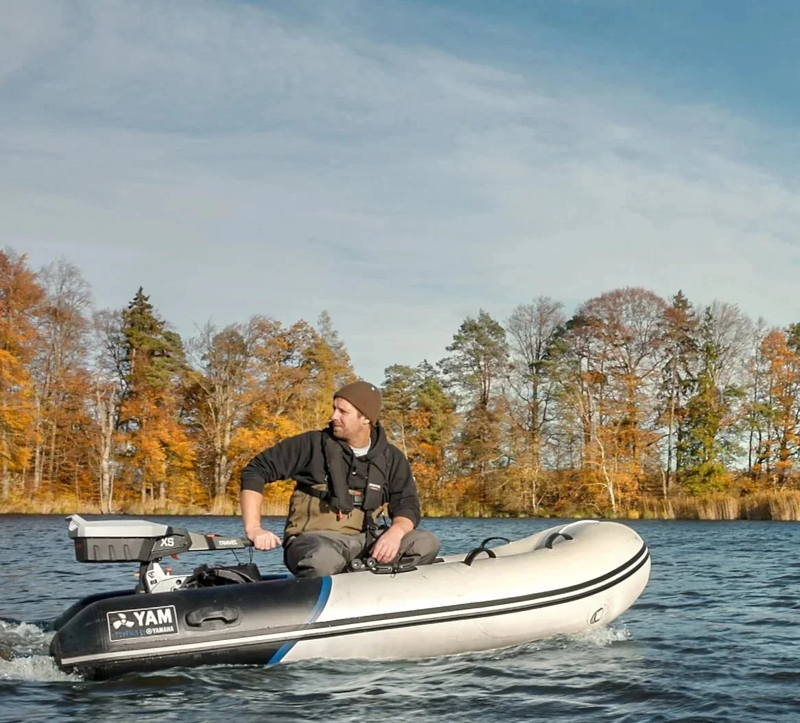 Man wearing a beanie and life jacket sitting on an inflatable boat on a lake with autumn trees in the background.