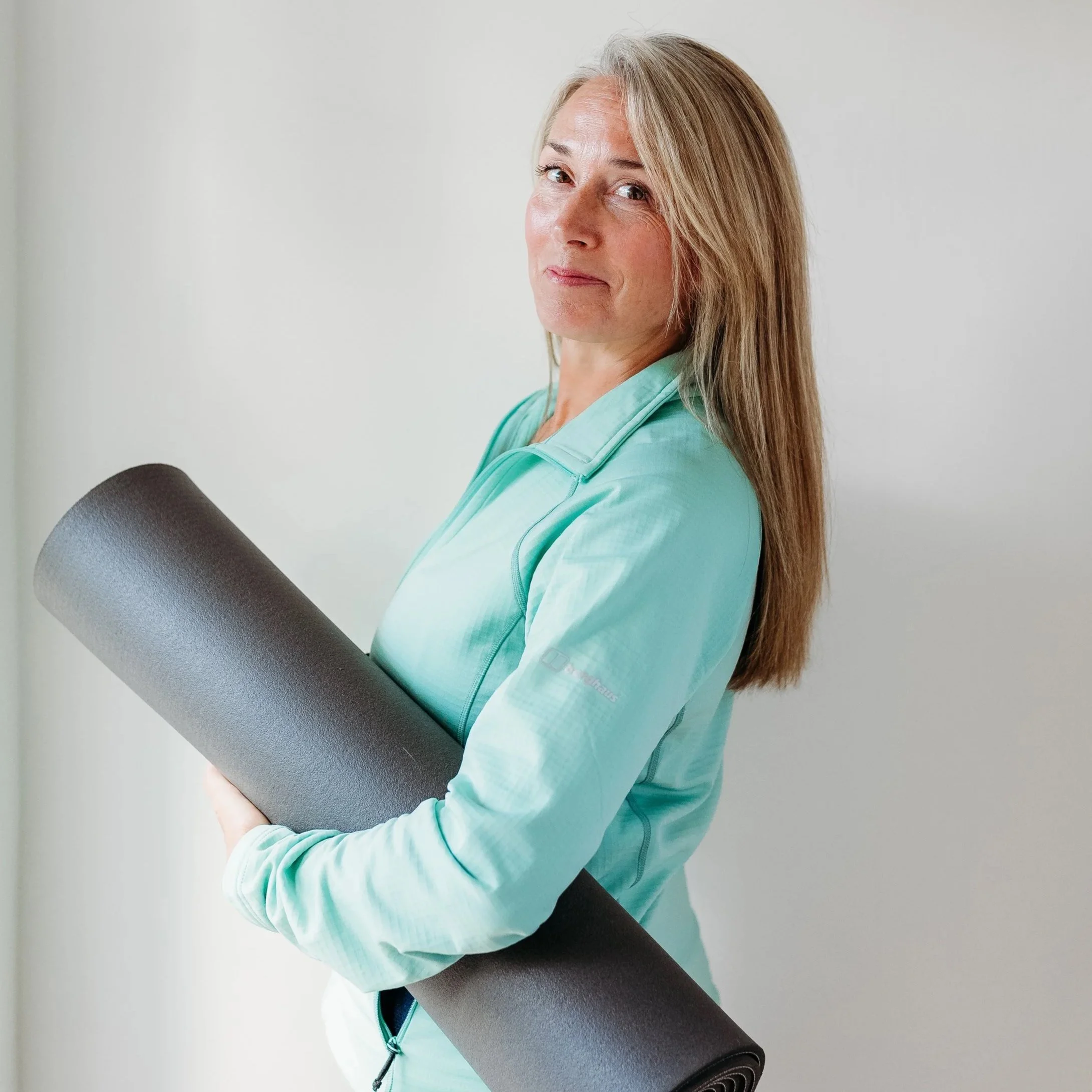Louise from the healing mat is  holding a black yoga mat, wearing a light green athletic jacket, with long blonde hair and a slight smile, standing against a plain white wall. Louise talks here about the workshops she offered with restorative yoga.