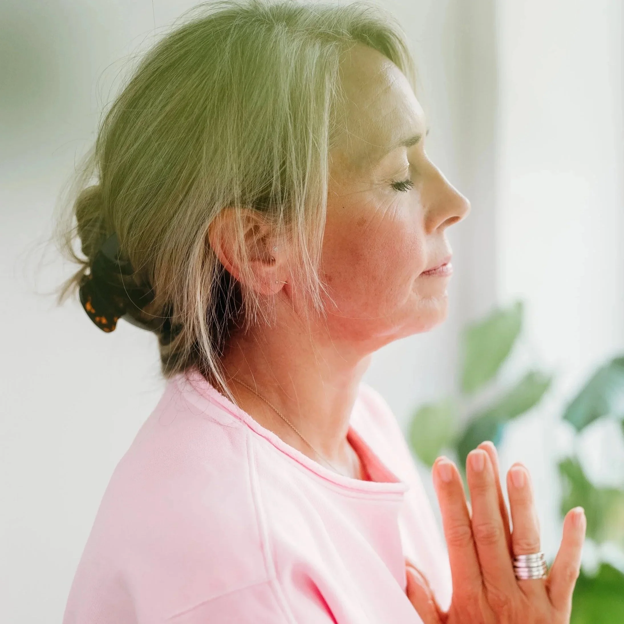 Louise from the healing mat has eyes closed, and hands pressed together near her chest, practicing meditation or prayer indoors near a window with natural light. Louise talks about her yoga classes and what she offered either in person or online.
