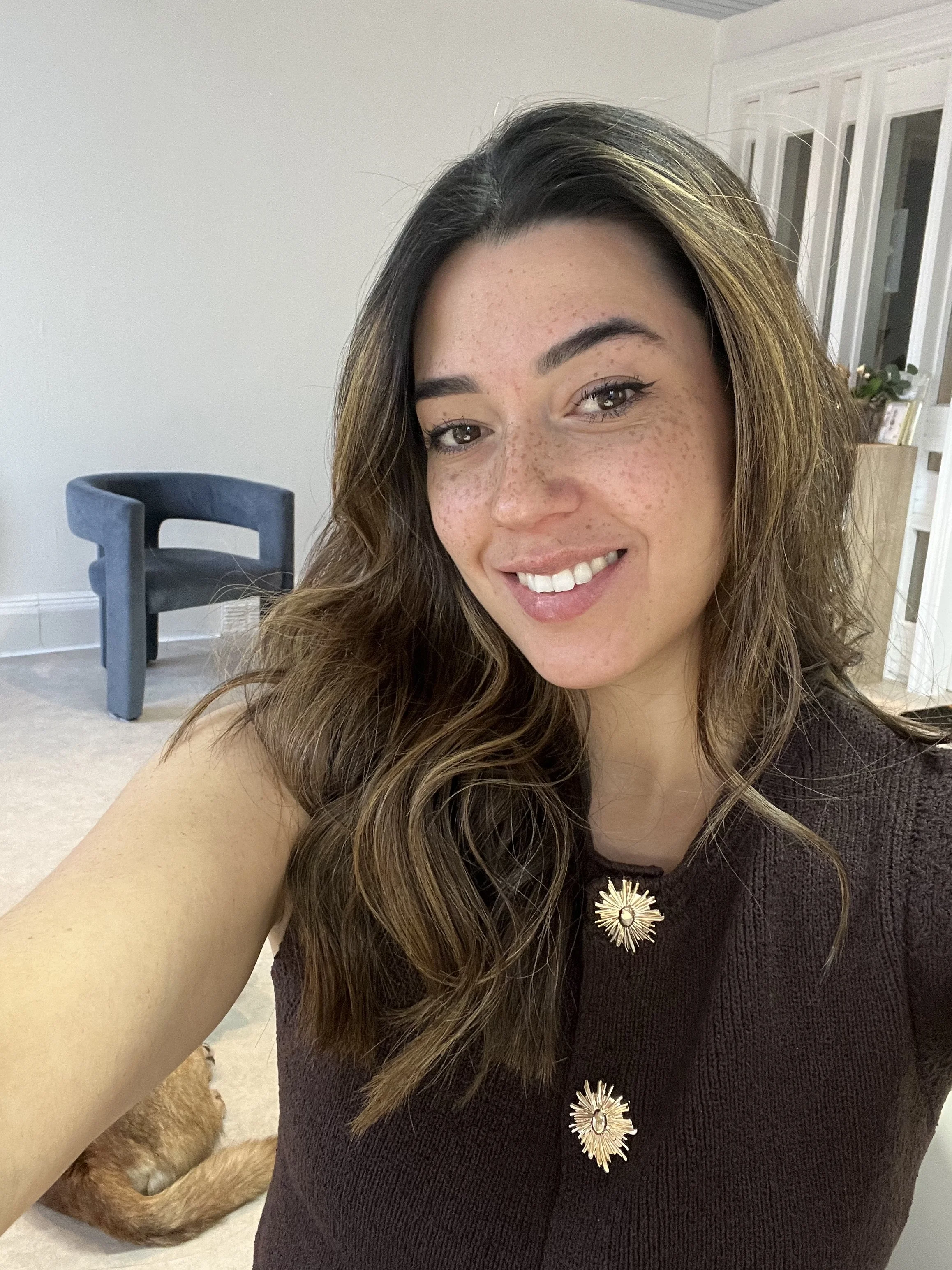 A woman with wavy brown hair and freckles taking a selfie in a well-lit room, smiling. She is wearing a dark top with decorative gold buttons. In the background, there is a blue armchair, white walls, a window with blinds, and part of a dog's tail is visible at the bottom of the image.