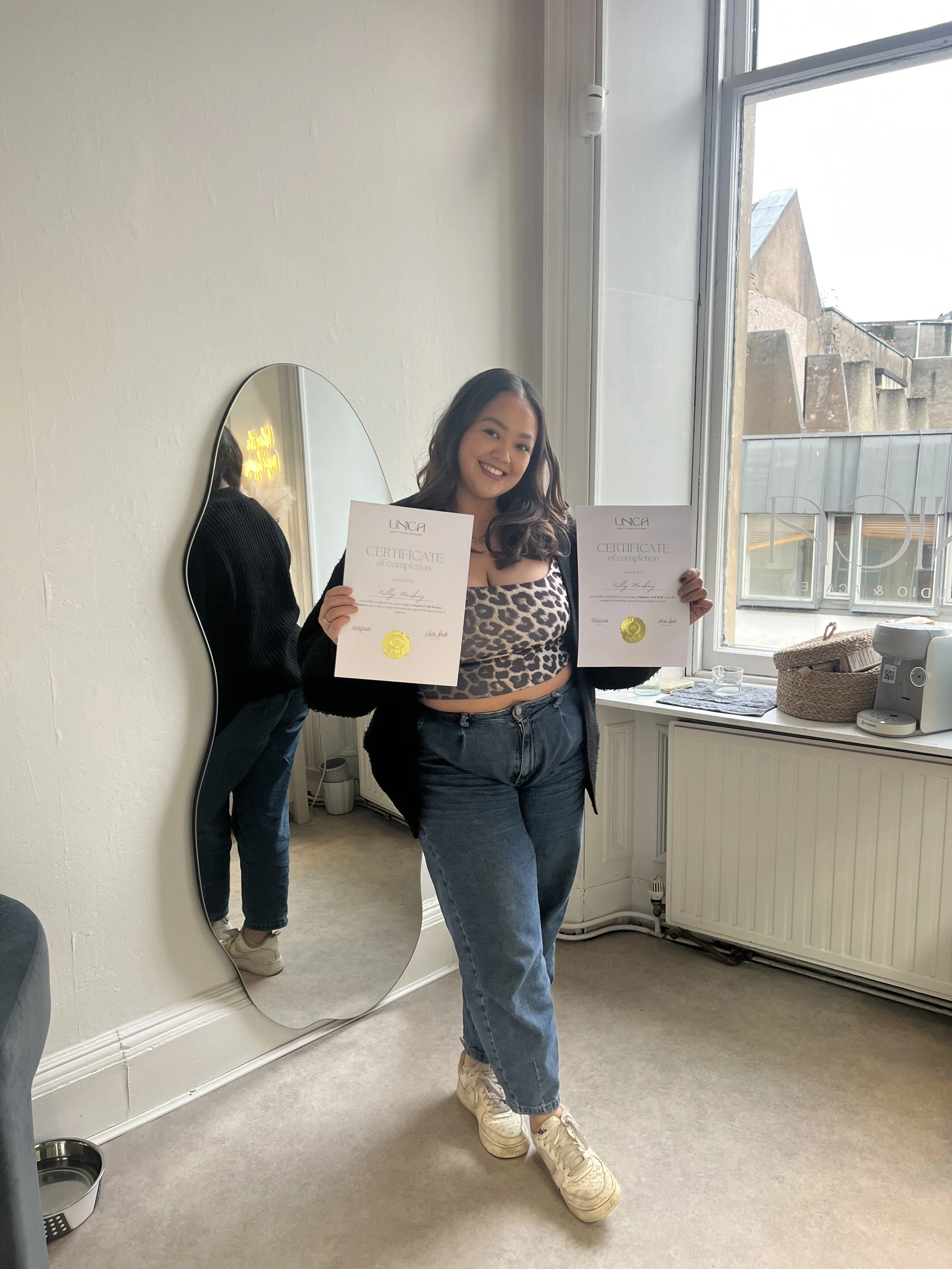 A woman standing inside, holding two certificates, smiling, near a window and mirror, with casual clothing, beige shoes, and dark jeans.
