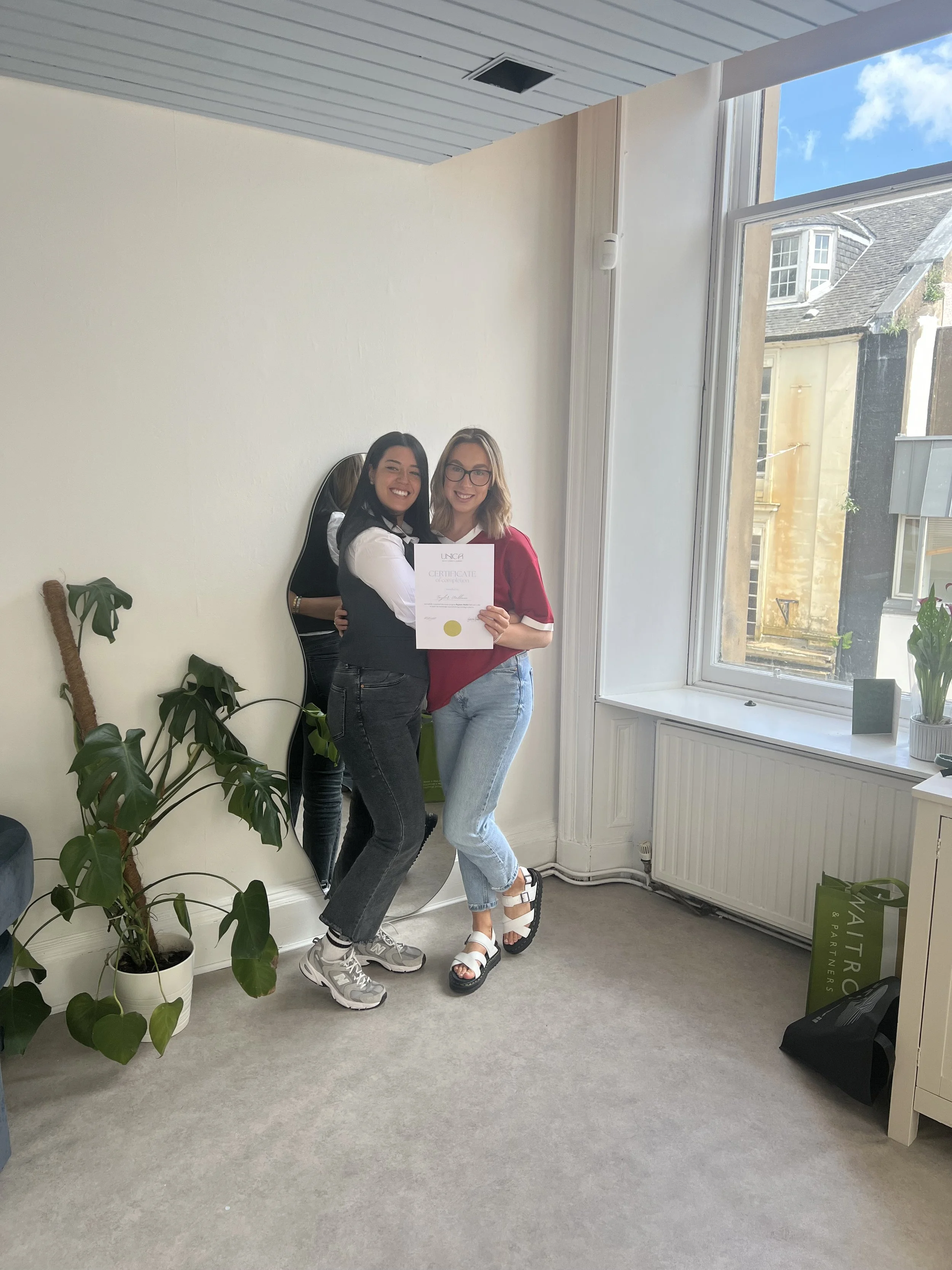 Two women standing together indoors, holding a certificate, smiling, near a large window with a view of neighboring buildings and blue sky, surrounded by potted plants and a full-length mirror.
