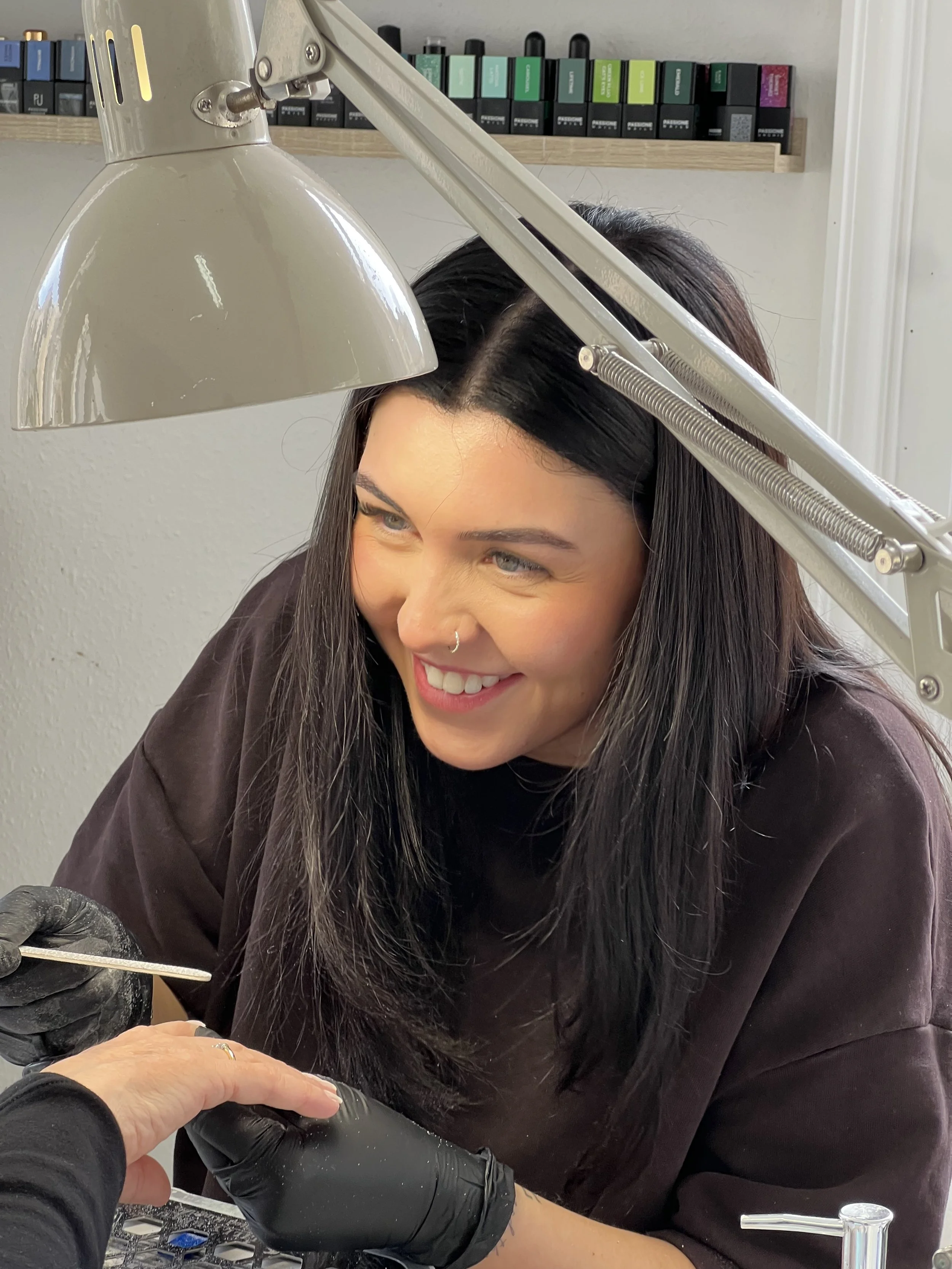A woman working on a tattoo with a tattoo needle, smiling and wearing black gloves, under a table lamp in a studio.