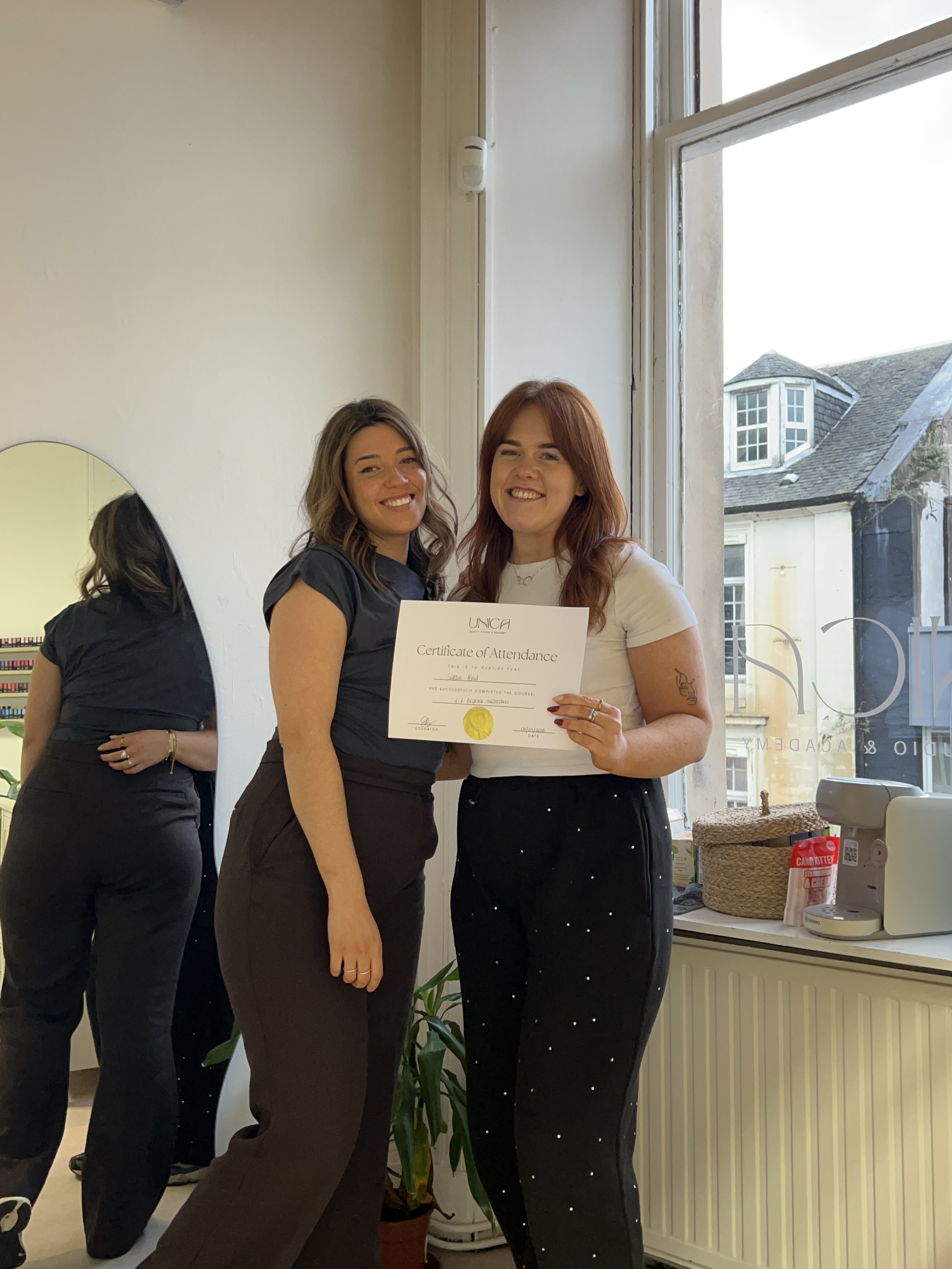 Two women smiling, one holding a certificate of attendance, standing near a window in a room with a mirror and houseplants.