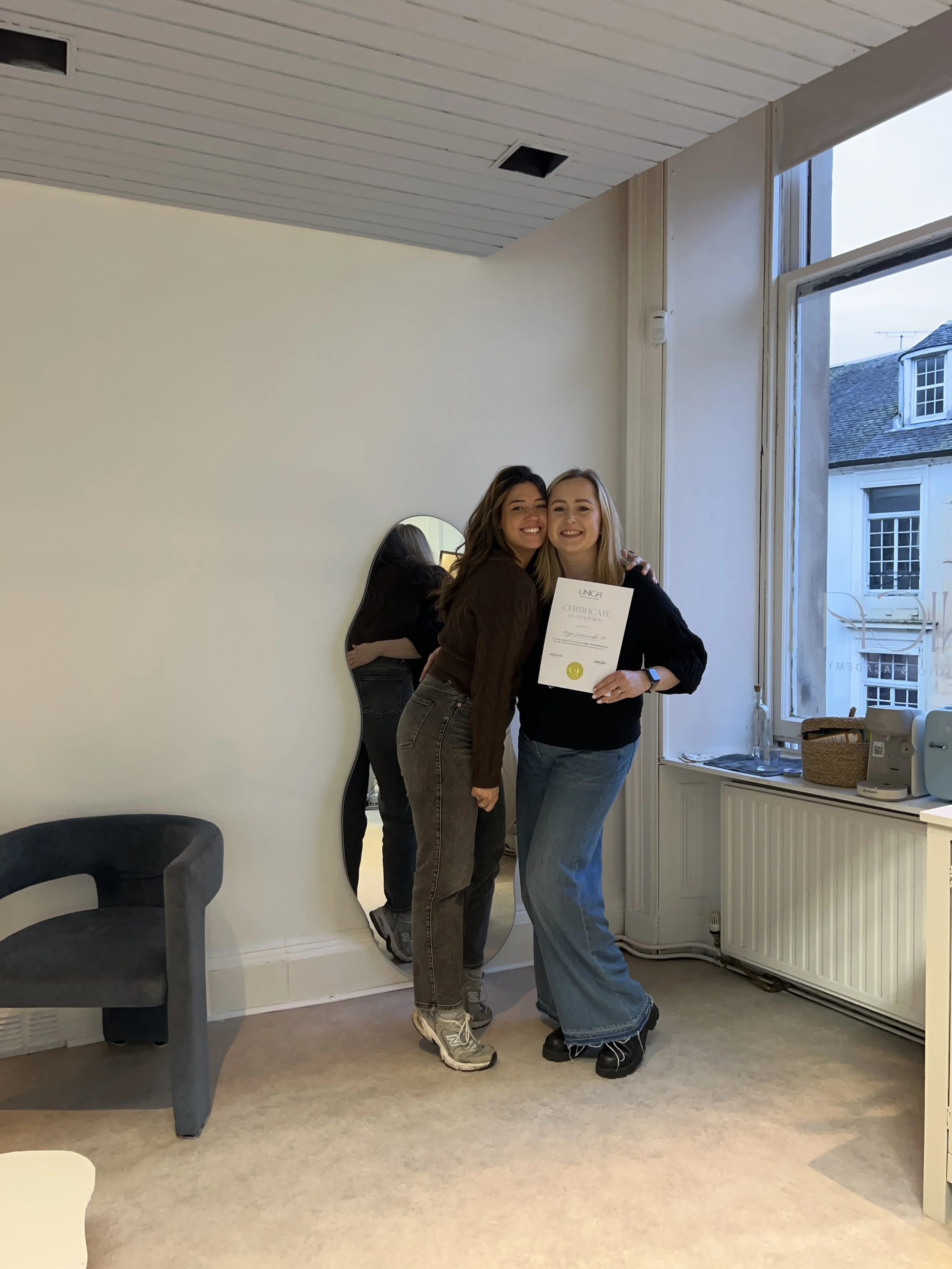 Two women smiling and hugging, one holding a certificate, standing near a window in a room with beige carpet and minimal decor.