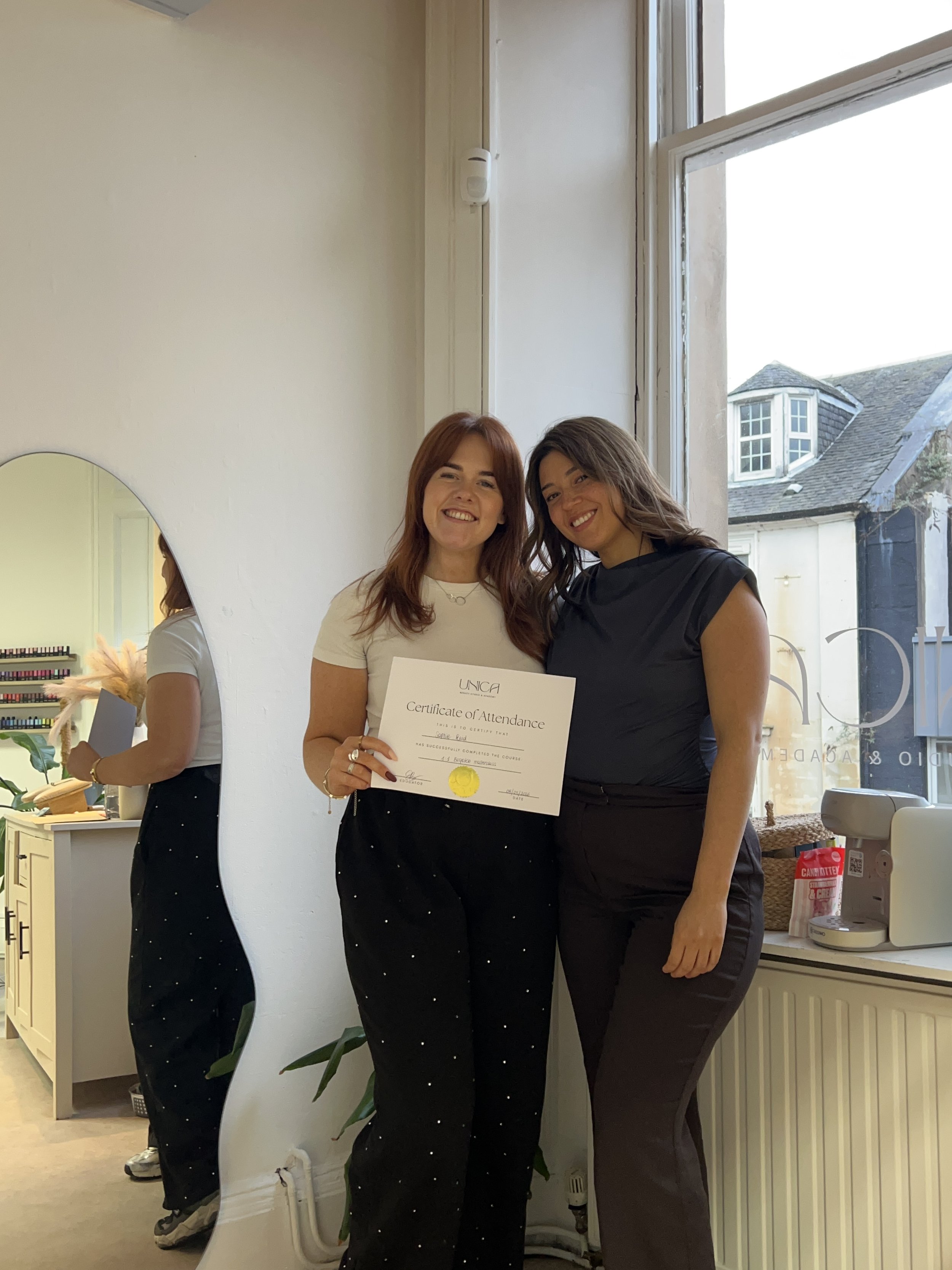 Two women standing next to each other smiling, one holding a certificate of attendance, in a well-lit indoor space with a window showing a house outside.
