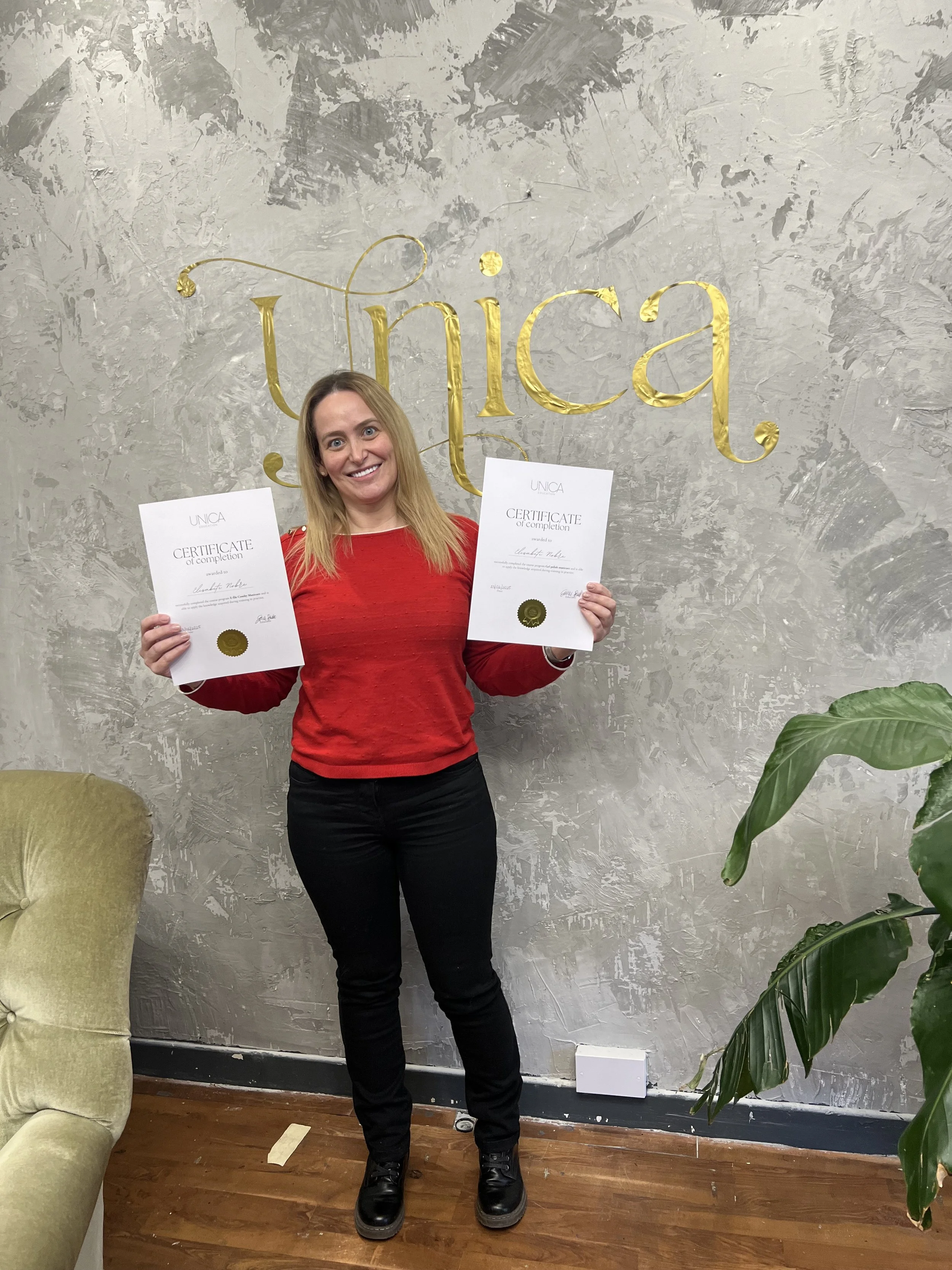 Woman in red sweater holding two certificates smiling in front of a wall with the word 'Unica' in gold letters, green plant on the right, and a beige armchair on the left.