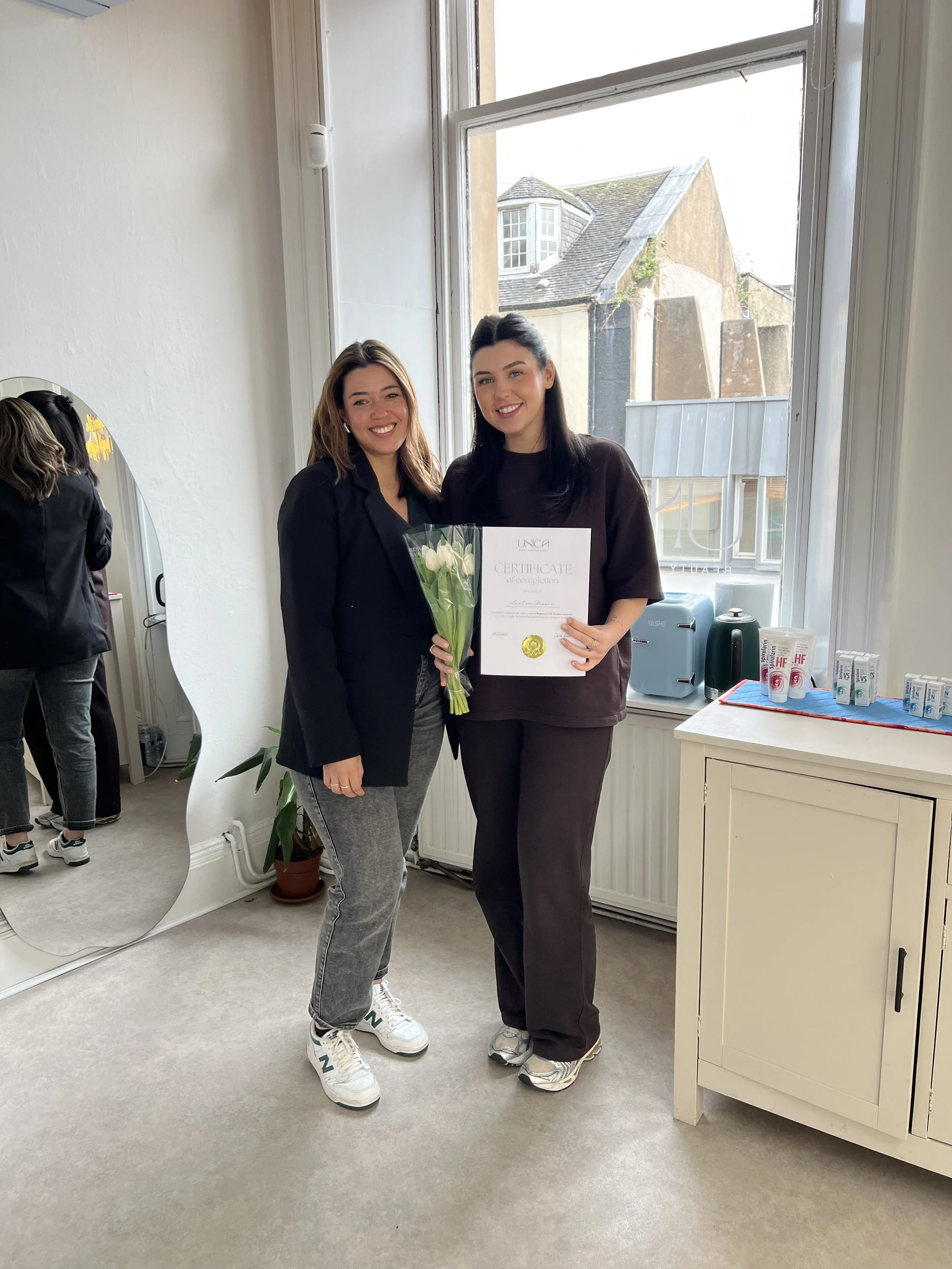 Two women smiling indoors, one holding a bouquet of white tulips and a certificate, with a window showing rooftops outside.