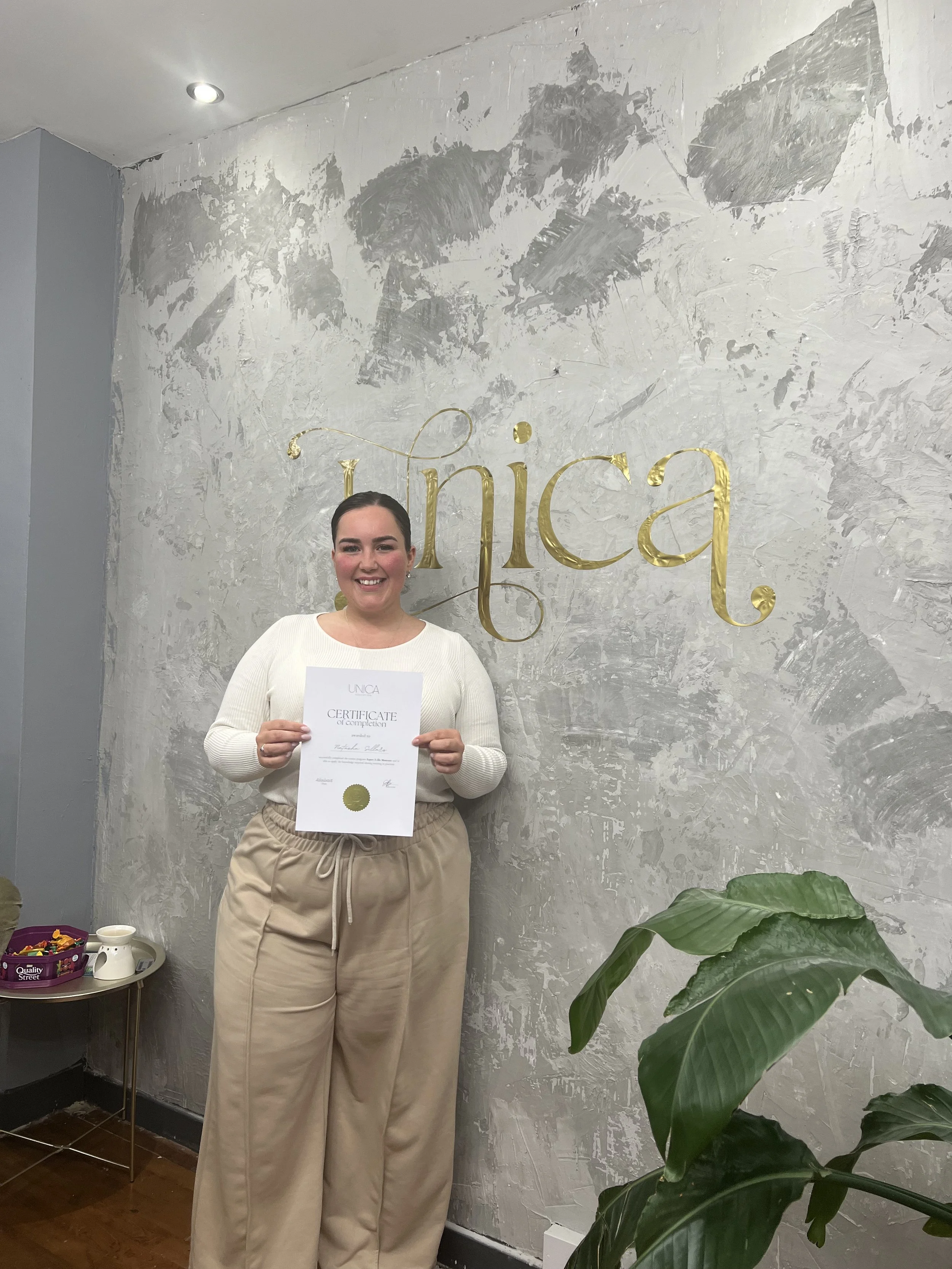 A woman holding a certificate while standing in front of a wall with the word 'Inica' in gold lettering. She is smiling and wearing a white sweater and beige pants.