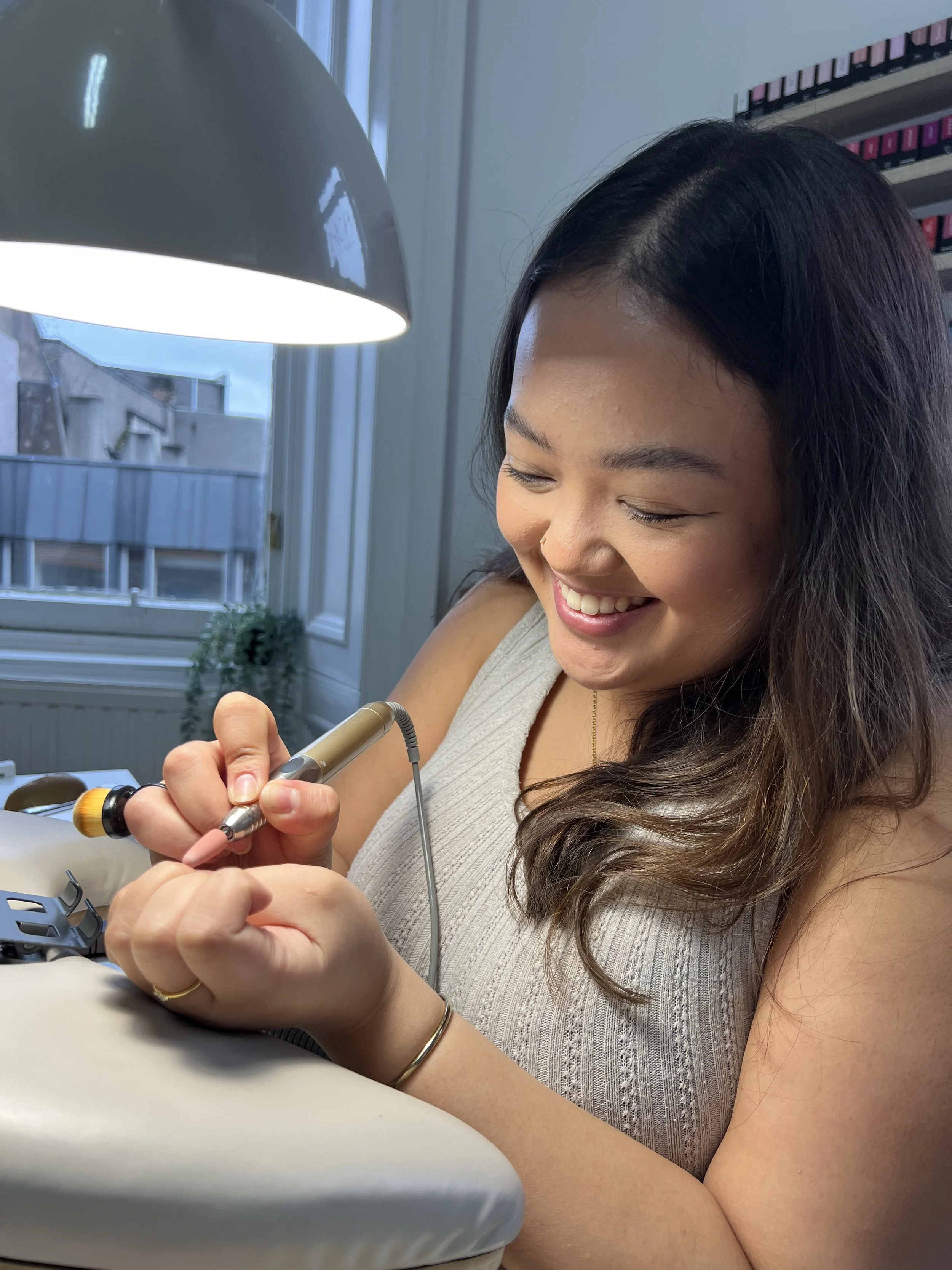 A woman smiling while working on a nail art device near a window with a lamp overhead.