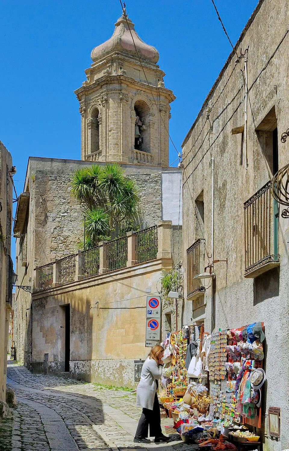 A cobbled stretch of Via Roma in Erice, with a bell tower, stone façades and a small souvenir display