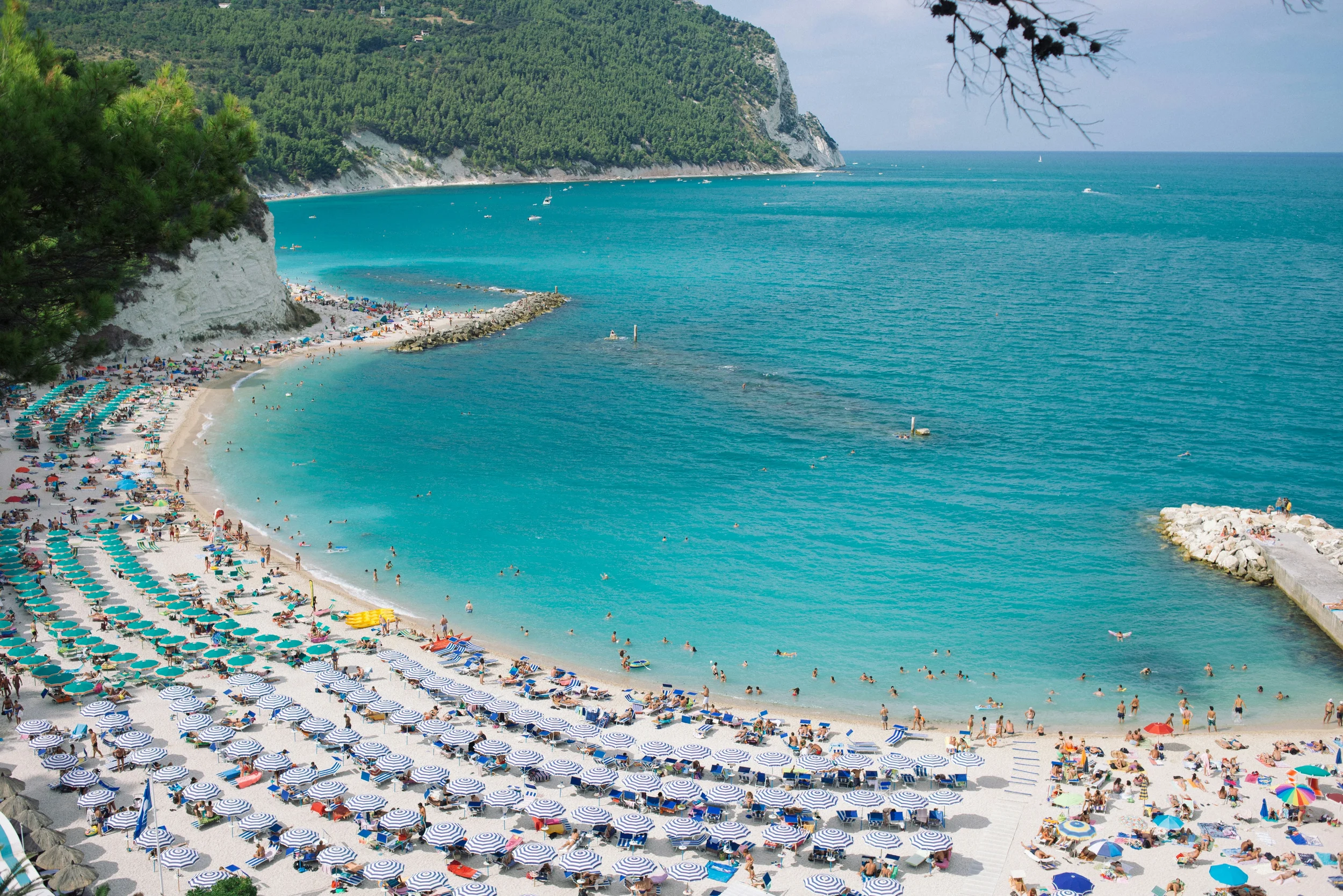 Curved beach with colourful umbrellas and turquoise water beneath wooded cliffs on the Conero Riviera in Le Marche, Italy