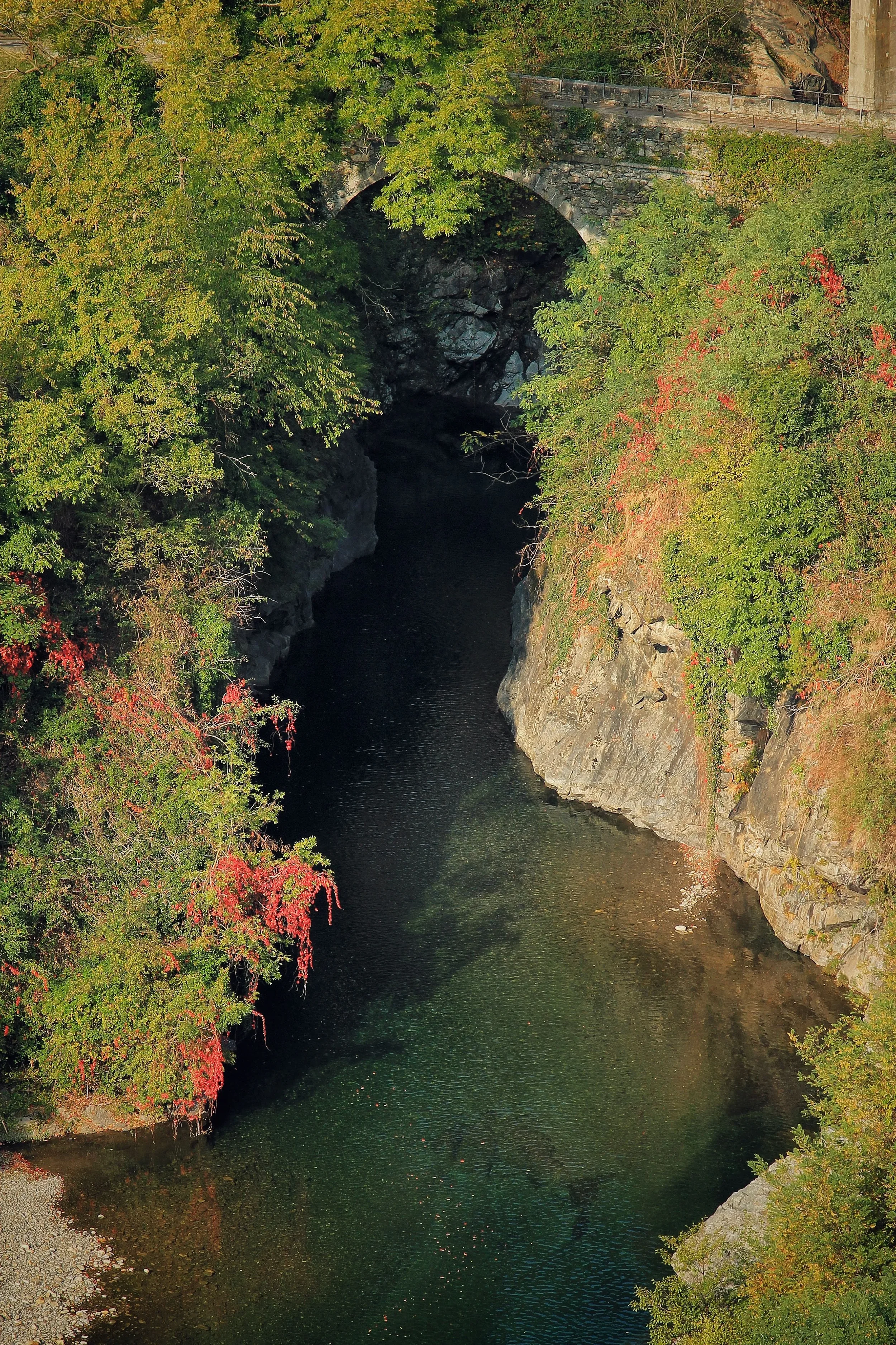 A stone arch bridge crosses a narrow rocky river gorge surrounded by autumn foliage.