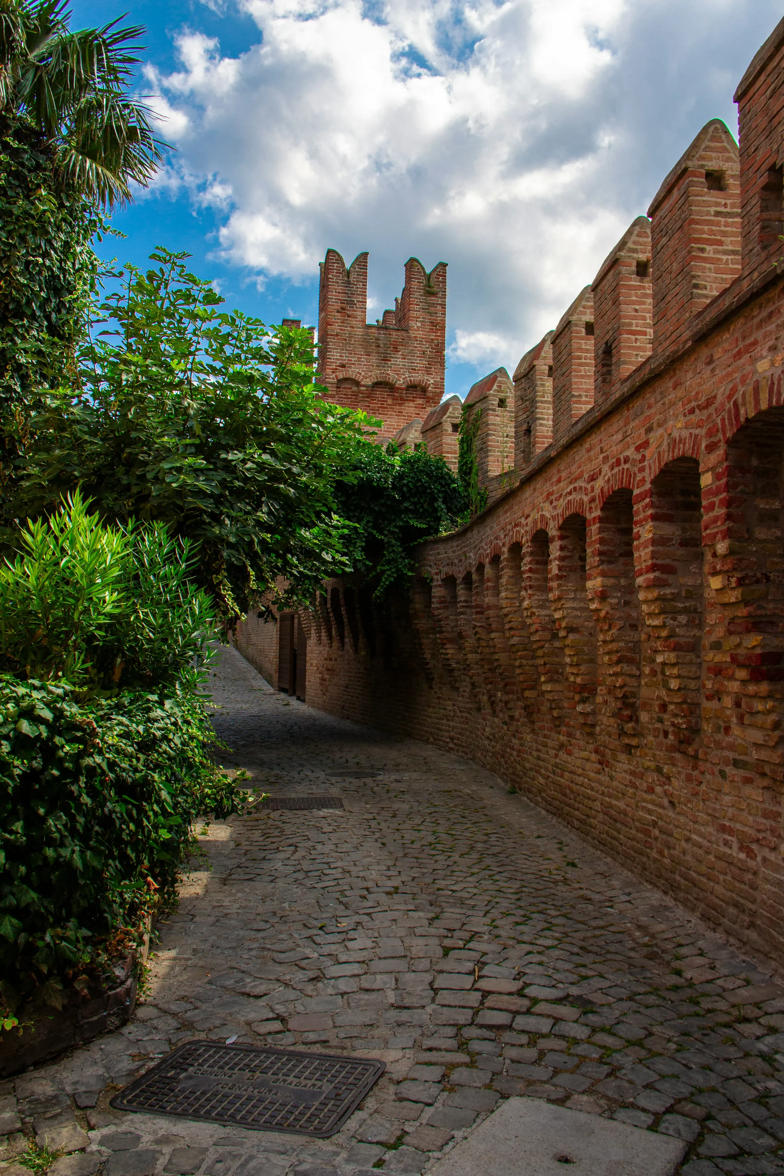 Cobbled path beside the crenellated brick walls of Gradara castle, bordered by green plants
