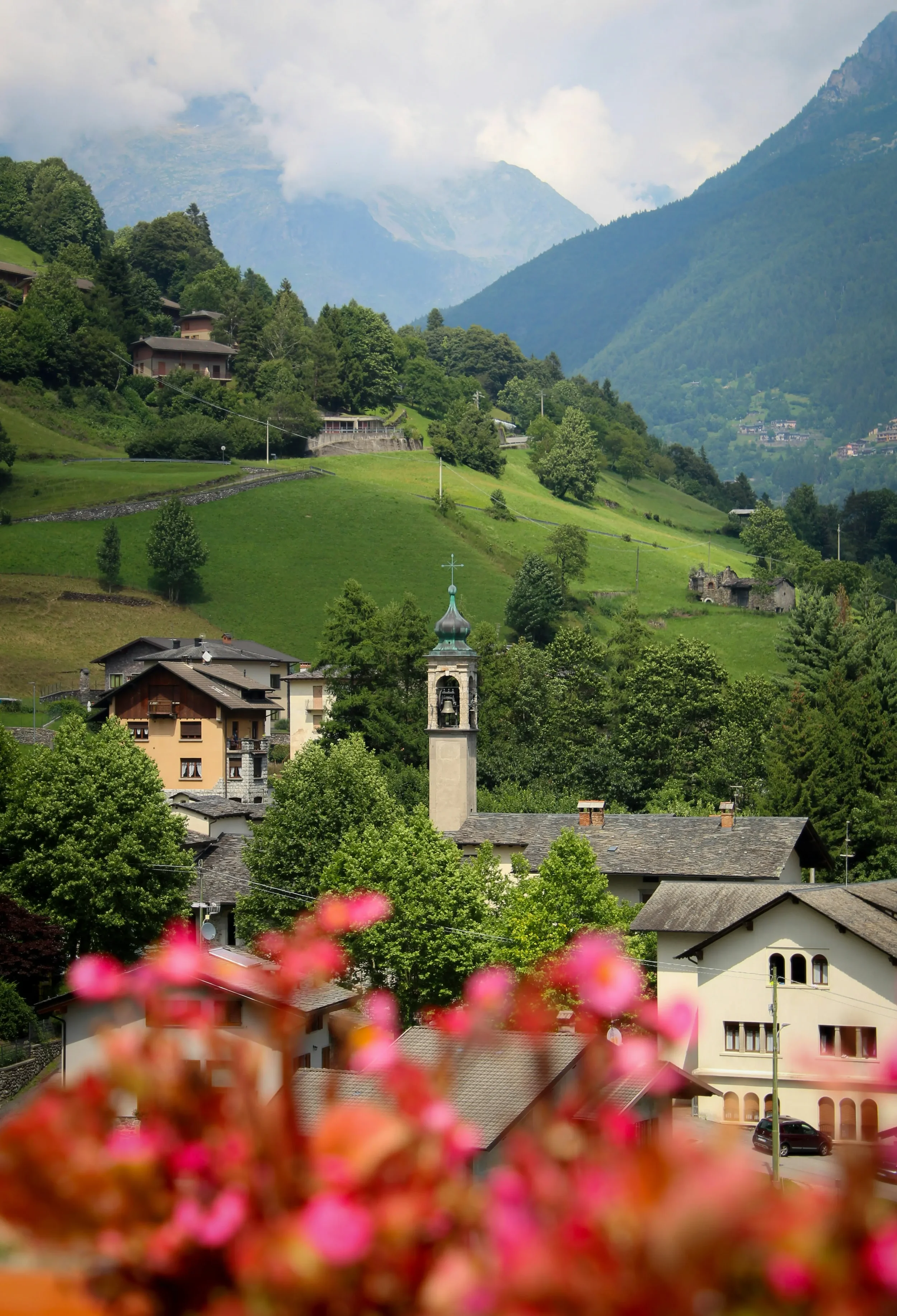A church bell tower among green hills and village houses, with flowers in the foreground.