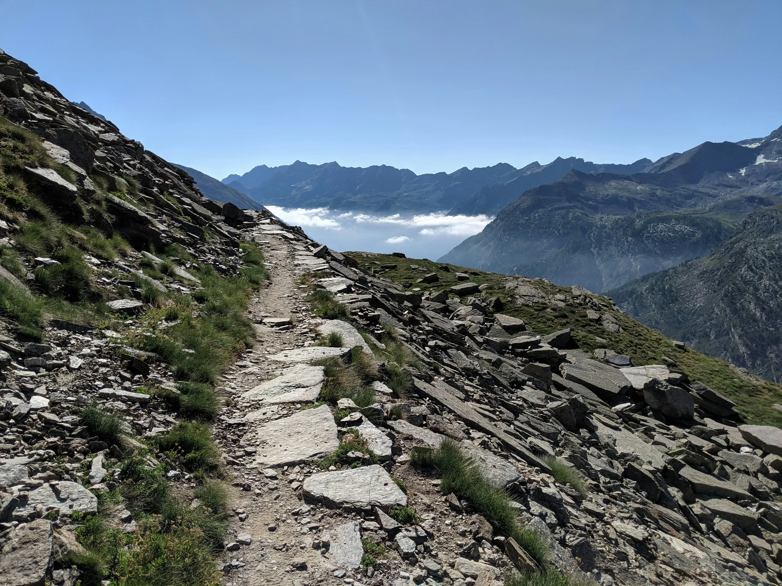 A rocky mountain footpath crossing a steep grassy slope above a cloud-filled valley.