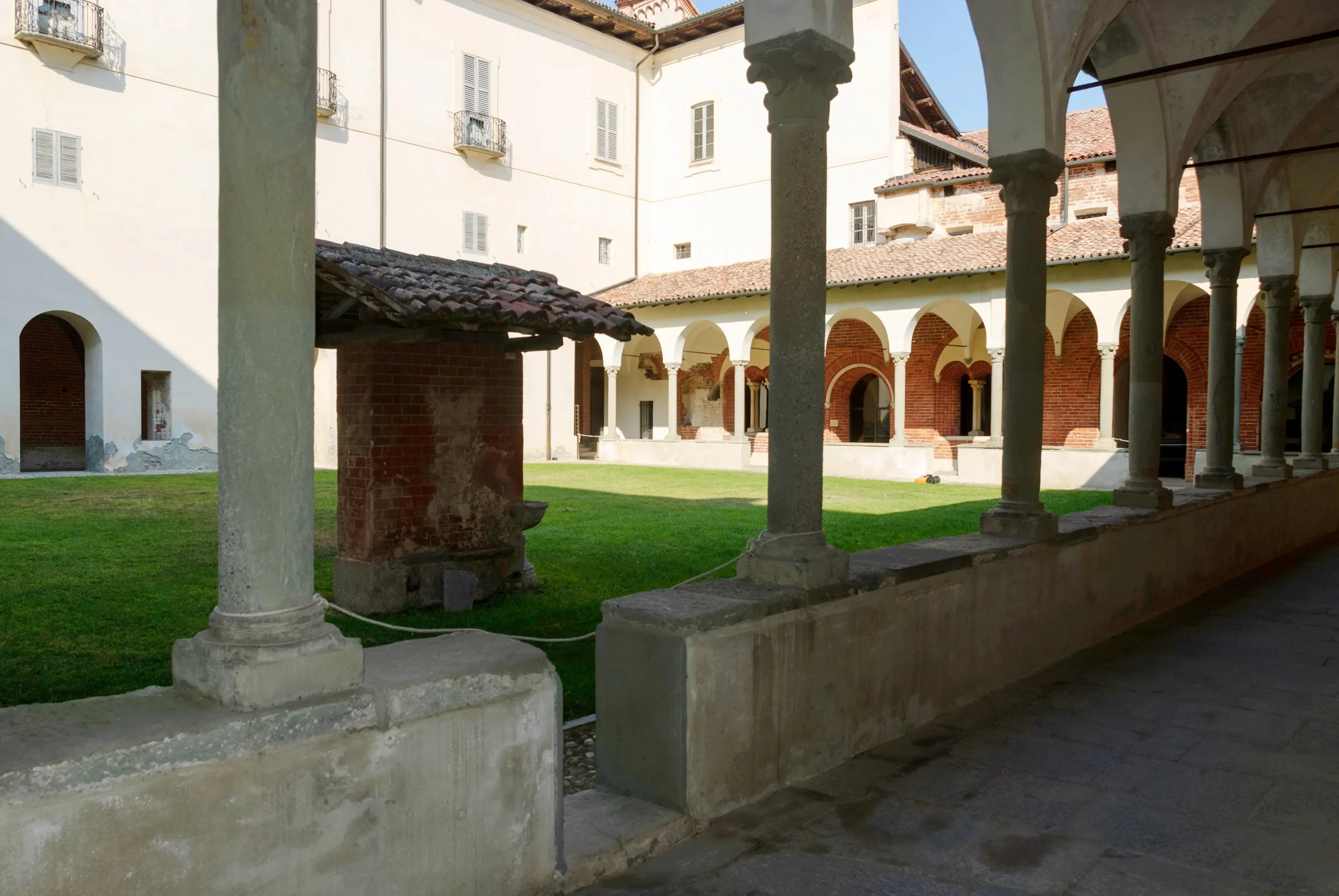 Arched cloister walk and covered brick well in the grassy courtyard of Morimondo Abbey.