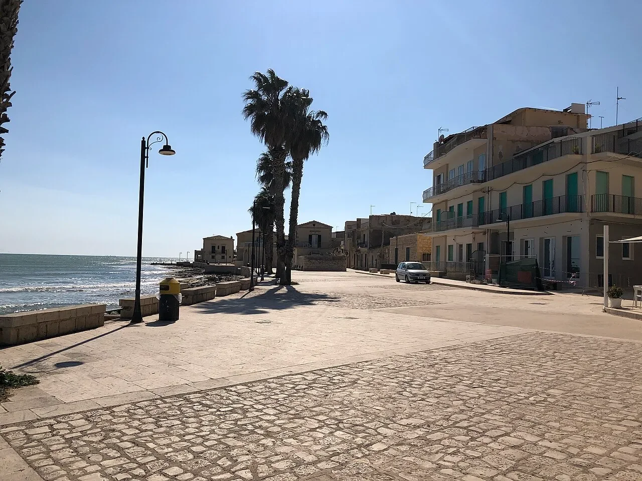 Sampieri’s seafront promenade, with palm trees, pale village buildings and waves along the shore