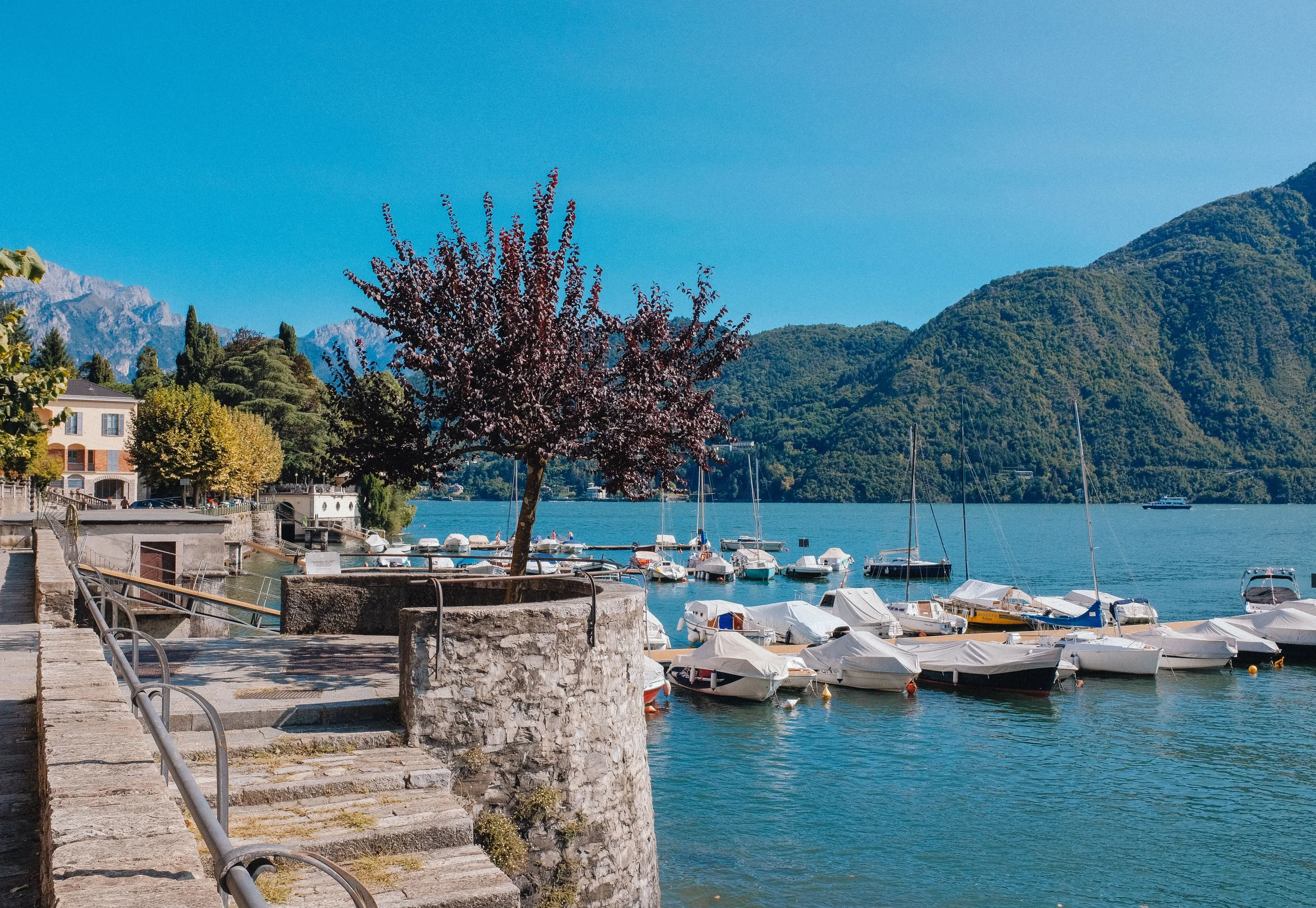 Small boats beside a stone lakeside promenade, with wooded mountains in the background