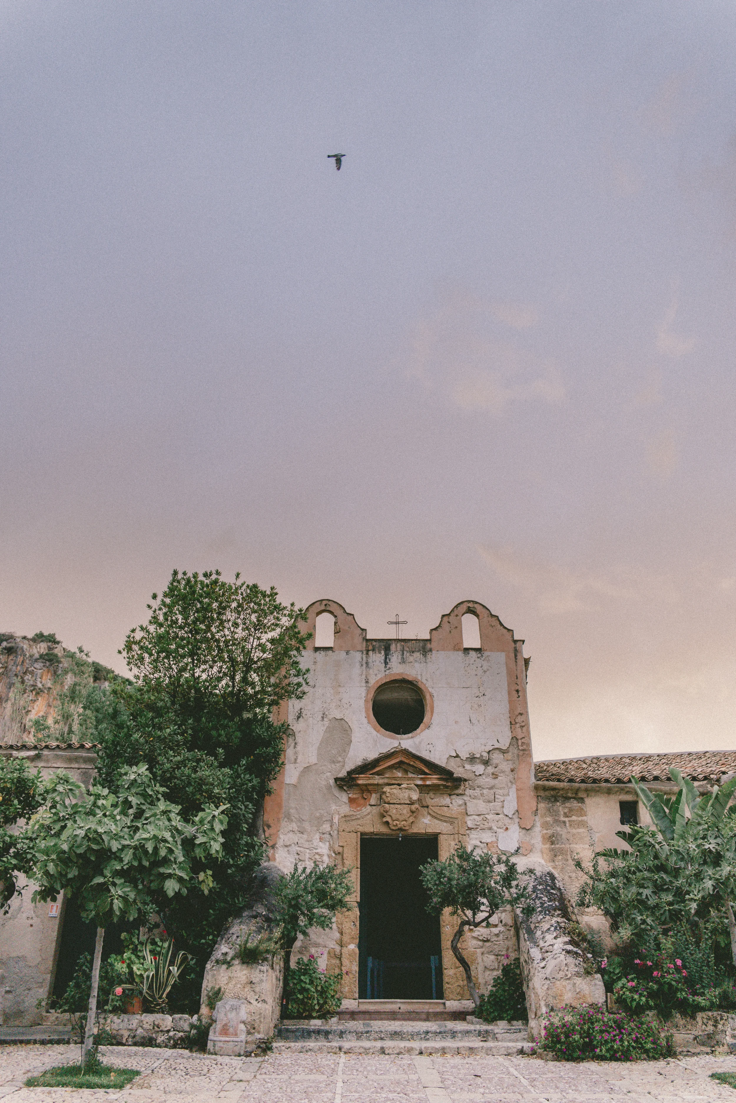 A weathered chapel facade with a round window, surrounded by trees and flowering plants in Scopello