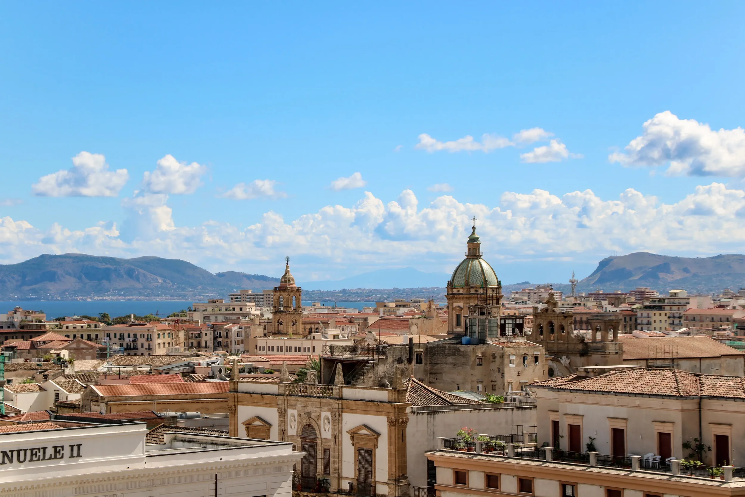 Panoramic view over a historic Sicilian town with church domes, terracotta rooftops and mountains in the background
