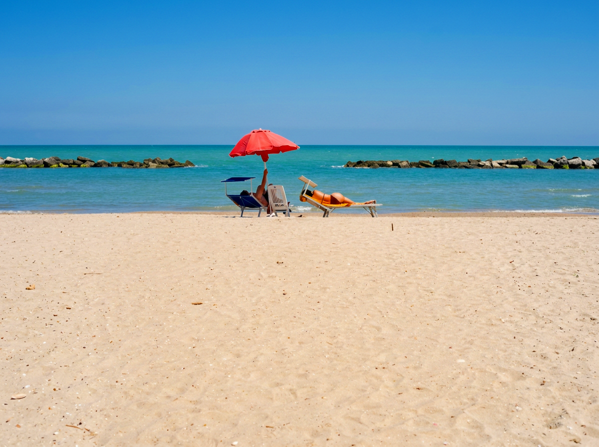 Person relaxing beneath a red parasol on the sandy beach of Grottammare, overlooking the blue Adriatic Sea in Le Marche, Italy