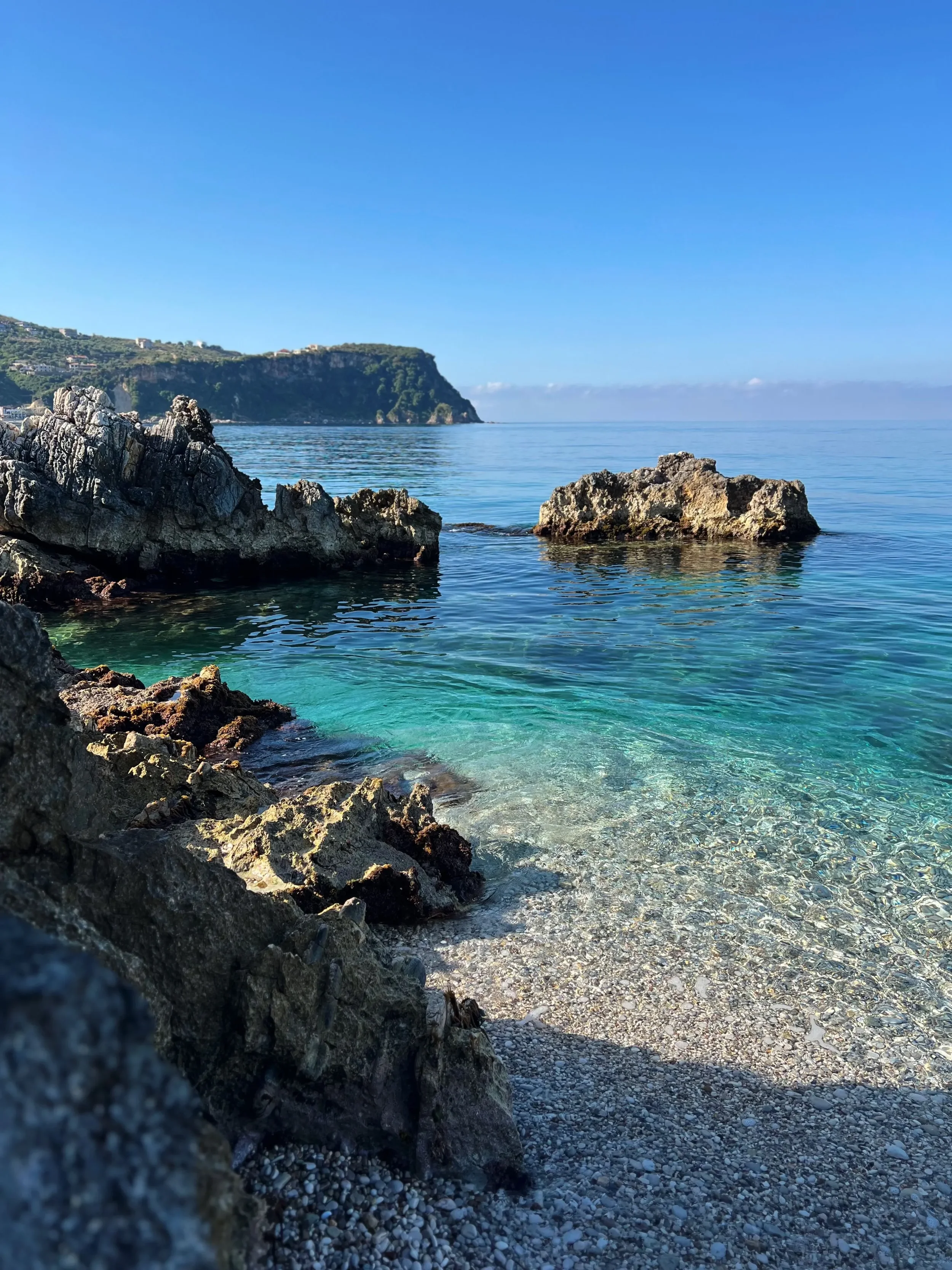 Clear blue-green sea washing over a small pebble beach, with dark shoreline rocks and an offshore rock beneath a blue sky.