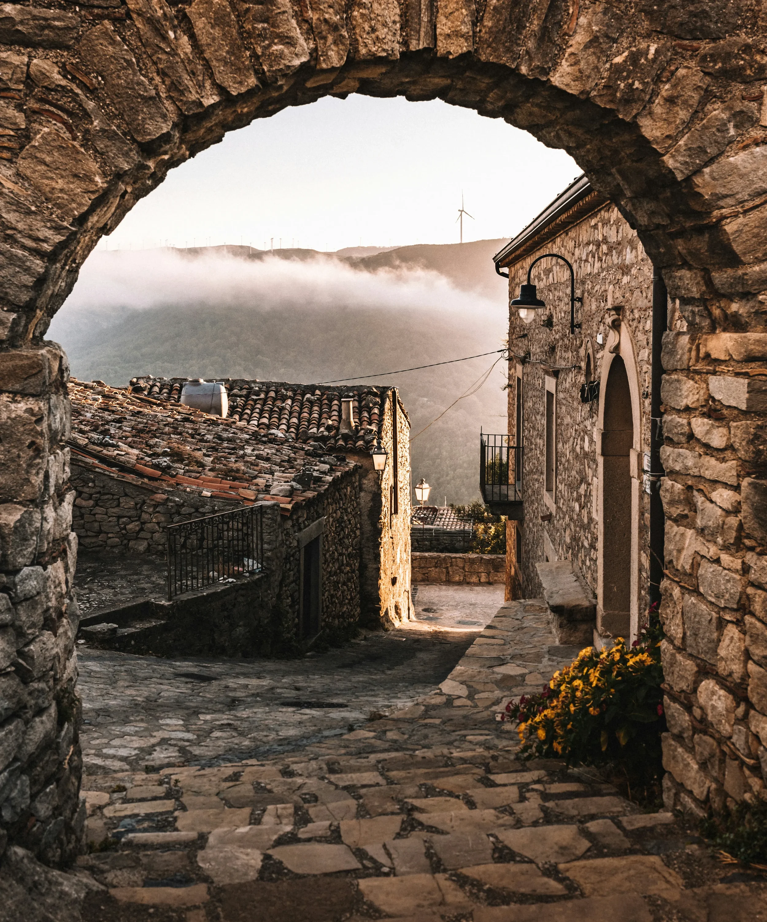 A stone arch framing a cobbled lane and tiled rooftops in Montalbano Elicona, with mist over the hills