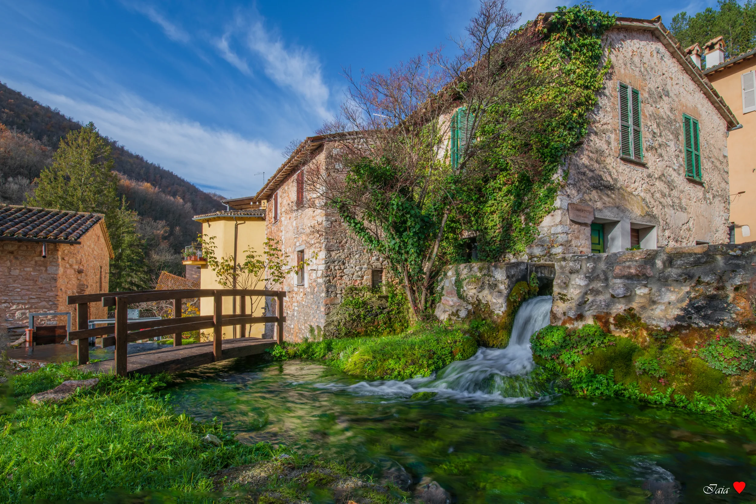 Clear spring water flowing beneath a wooden footbridge beside ivy-covered stone houses in Rasiglia