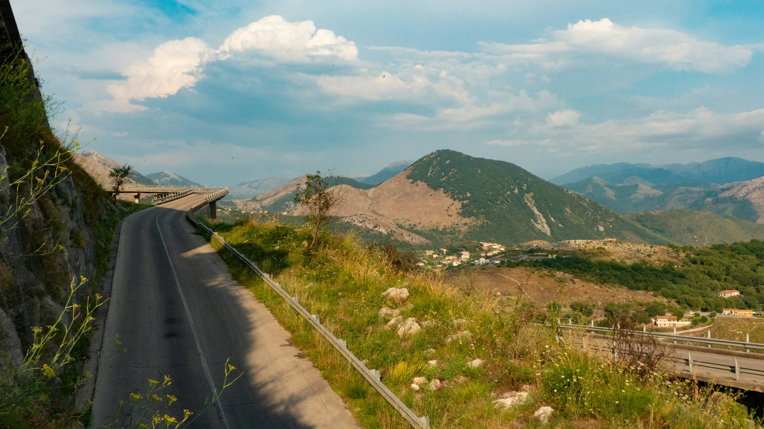 Winding mountain road through rugged hills and green countryside beneath a partly cloudy sky