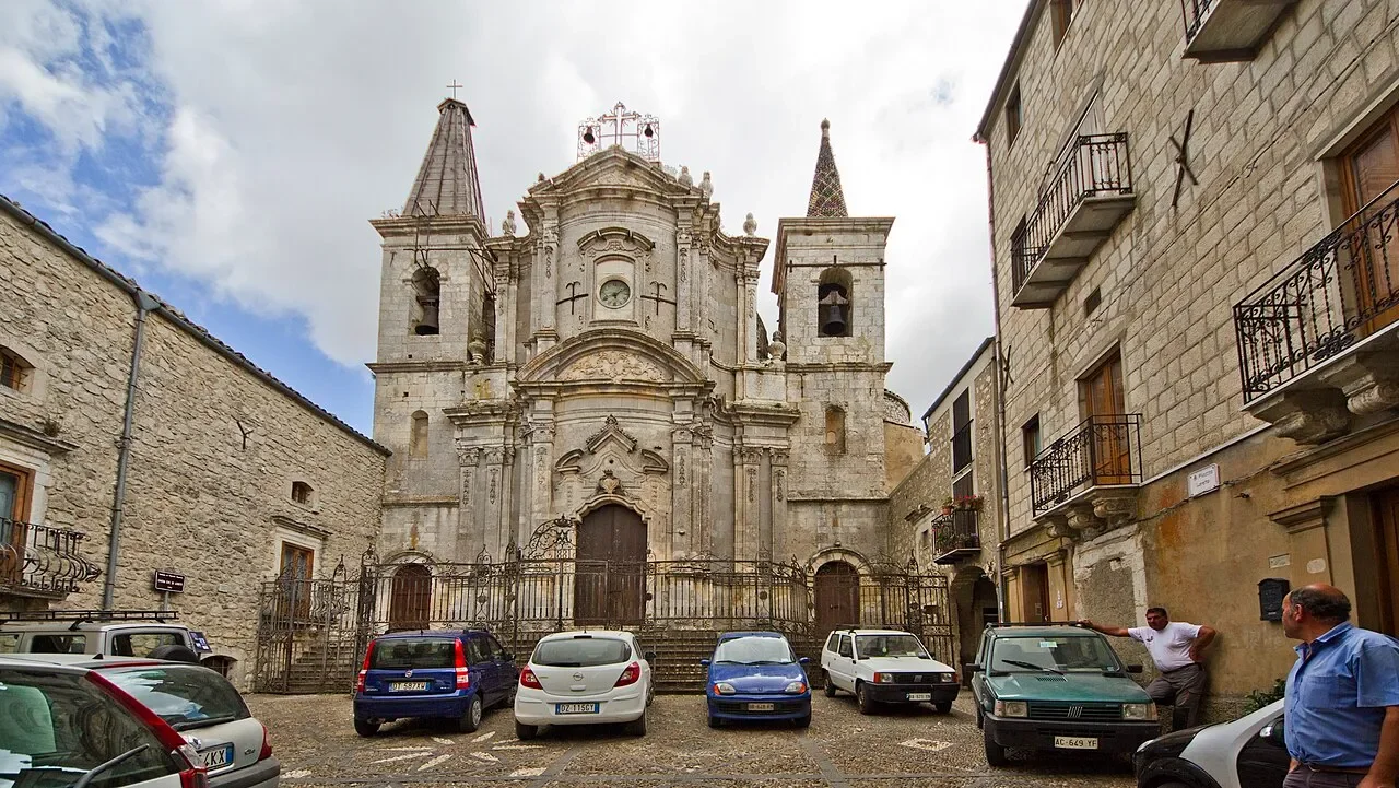 The curved facade and twin spires of Santa Maria di Loreto in Petralia Soprana, with parked cars in the square