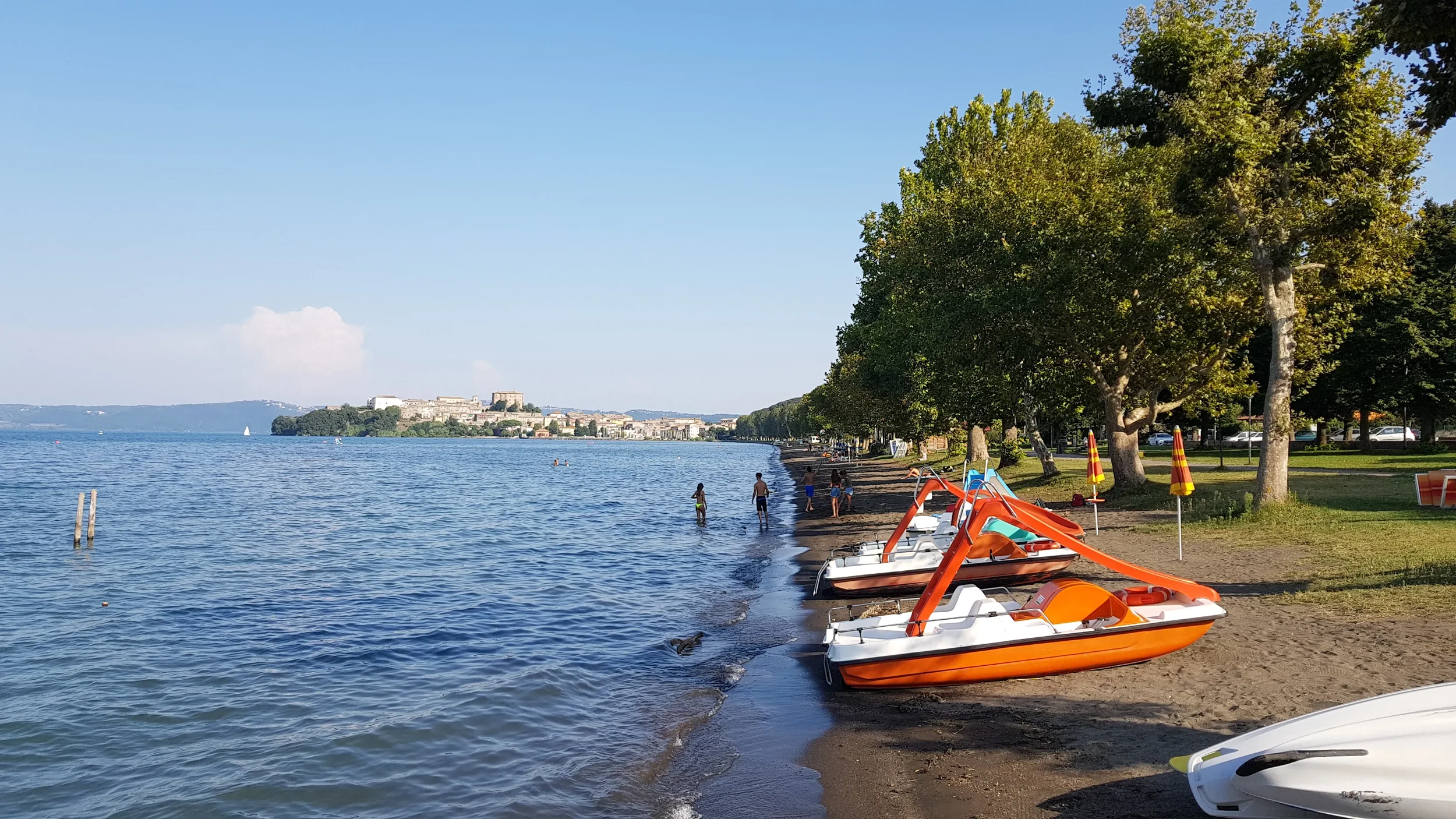 Pedal boats on the tree-lined shore of Lake Bolsena at Capodimonte, with the town across the bay.