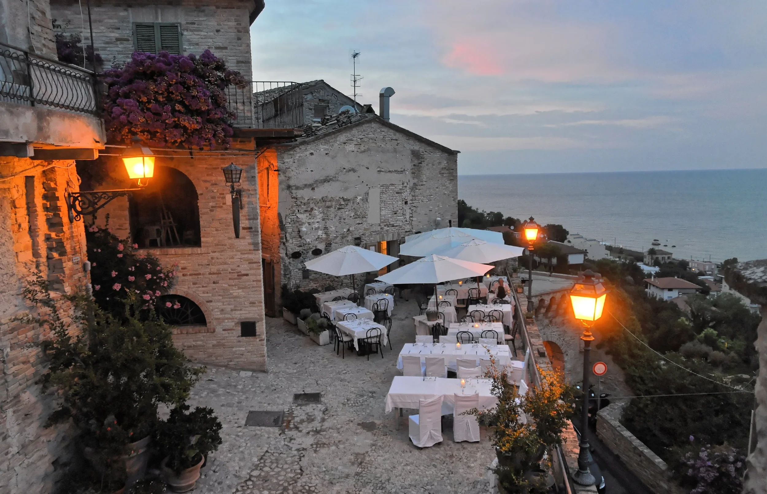 Lantern-lit restaurant terrace among brick buildings in Grottammare Alta, overlooking the Adriatic Sea at dusk