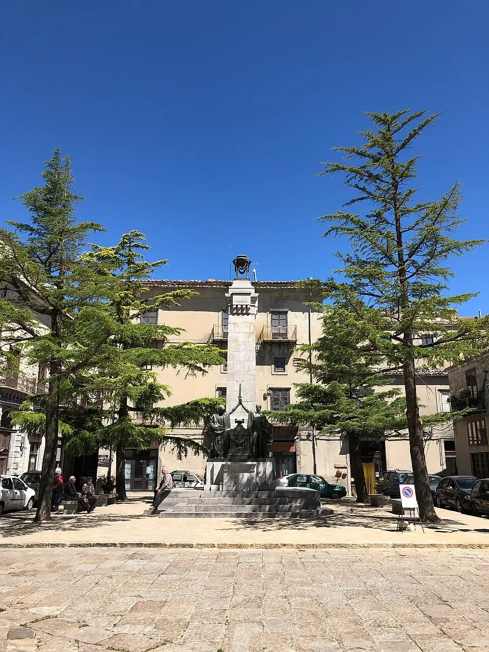The war memorial framed by tall trees in Piazza del Popolo, Petralia Soprana